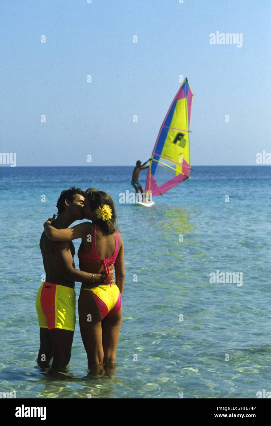 giovane capelli castani felice baciare coppia indietro vista armoniosa tuta bagnaria colorata con windsurf vacanze cielo blu mare sfondo Foto Stock