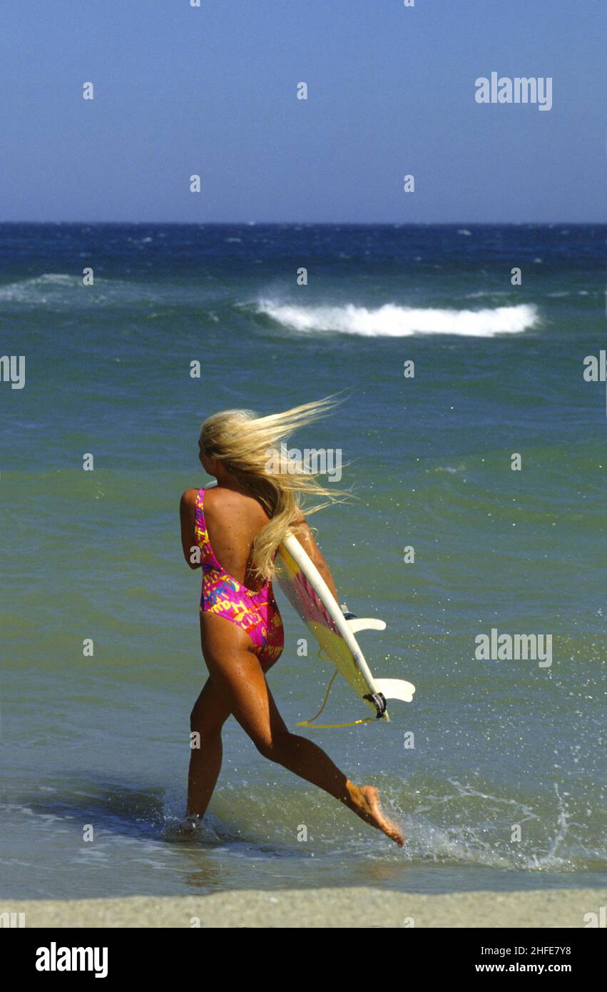 bellezza attraente capelli biondi sportivo retro silhouet profil correre in spiaggia con surf board spruzzi blu cielo mare sfondo Foto Stock
