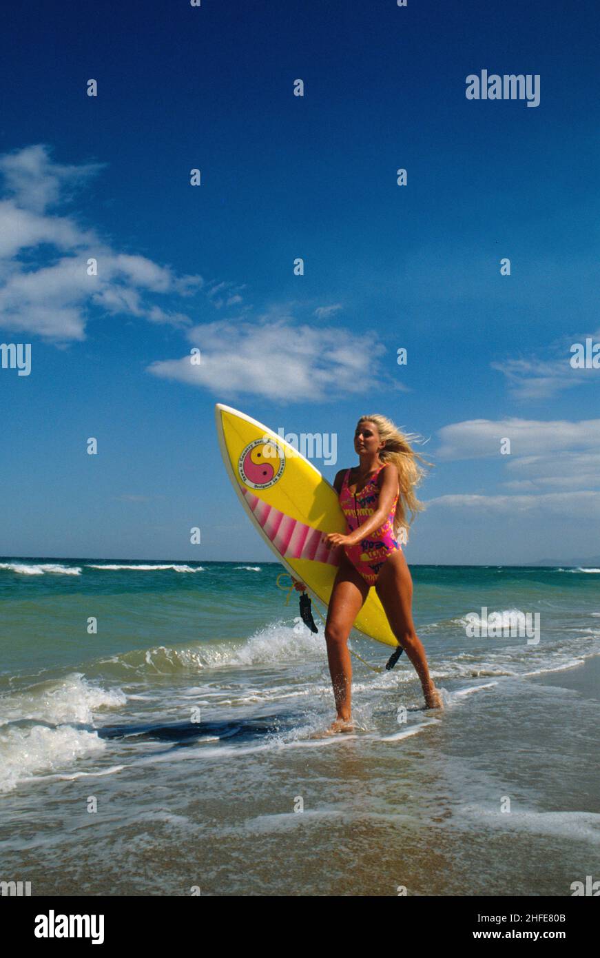 bellezza attraente capelli biondi sportivo retro silhouet profil correre in spiaggia con surf board spruzzi blu cielo mare sfondo Foto Stock