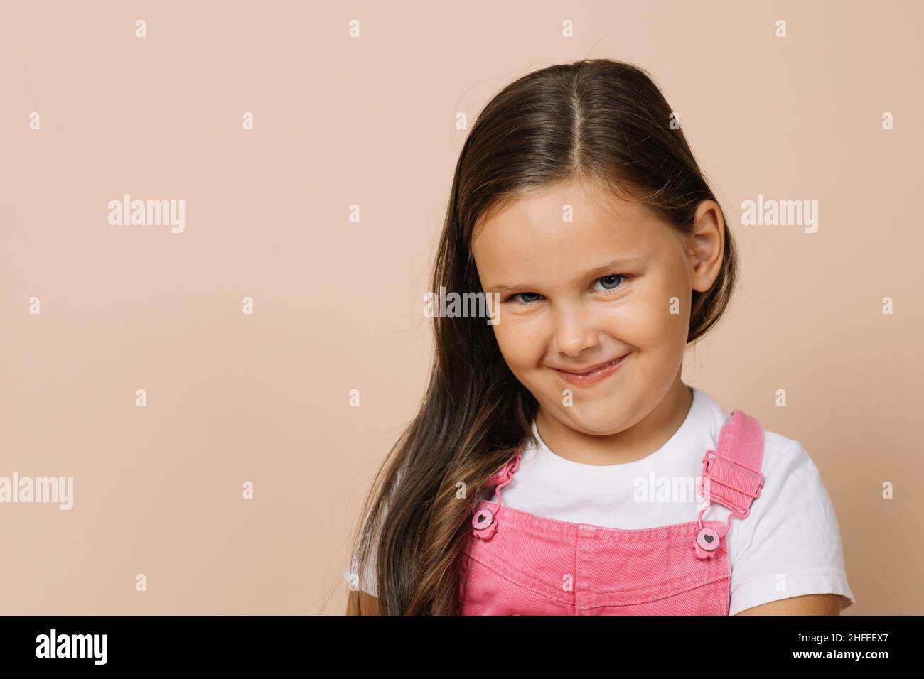 Primo piano ritratto di femmina capretto con occhi luminosi, sorriso umile e sguardo sullen guardando la macchina fotografica che indossa jumpsuit rosa brillante e t bianco Foto Stock
