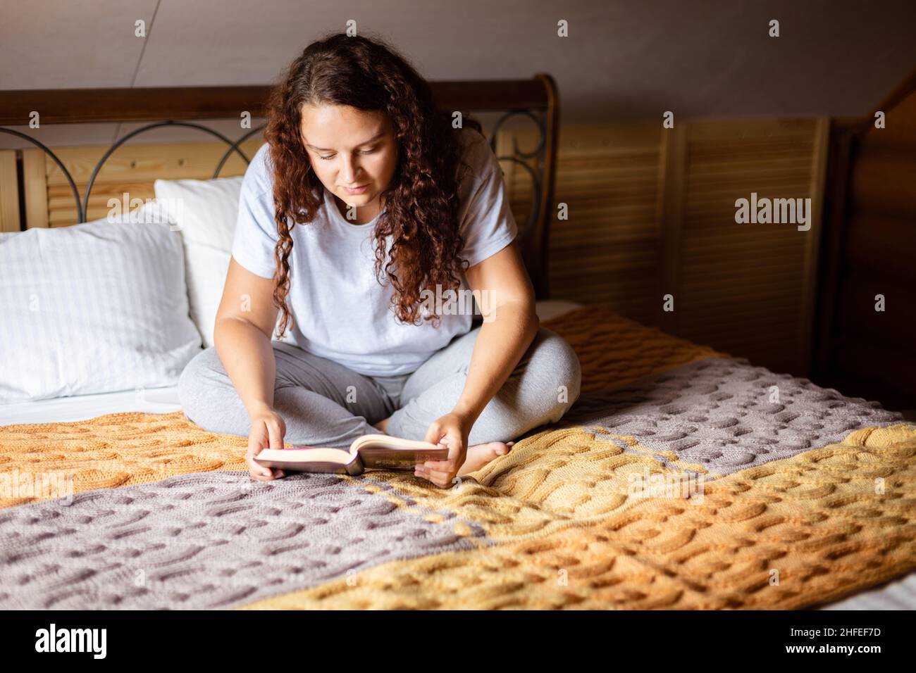 Ricci donna adulta seduta a gambe incrociate sul letto coperto con copriletto in spugna con libro in mani, lettura con grande fuoco indossando abiti a casa a. Foto Stock