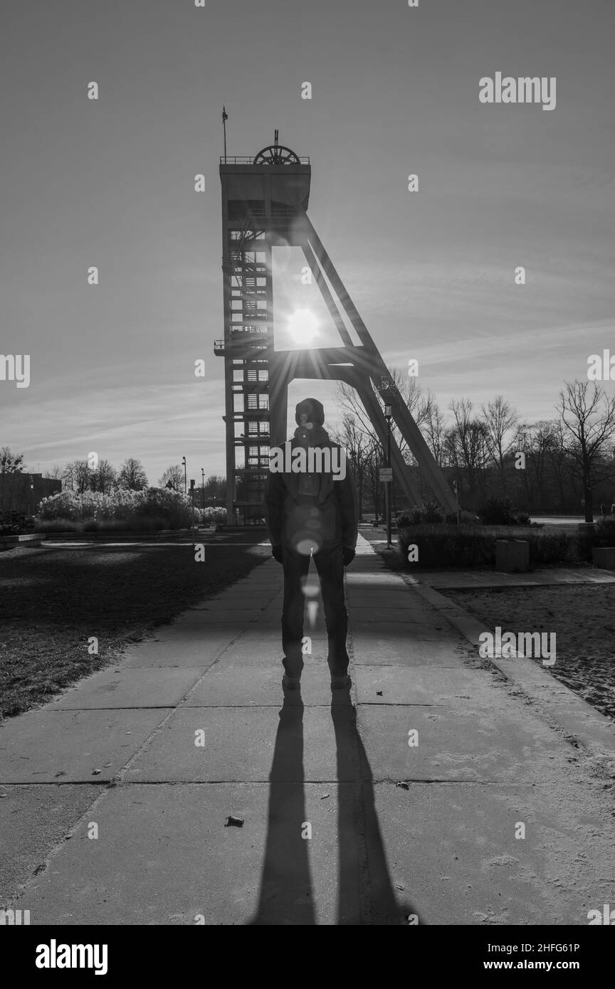 Un uomo in giacca invernale rossa che guarda i raggi del sole attraverso la struttura di un albero di miniera nel paesaggio della Slesia Foto Stock