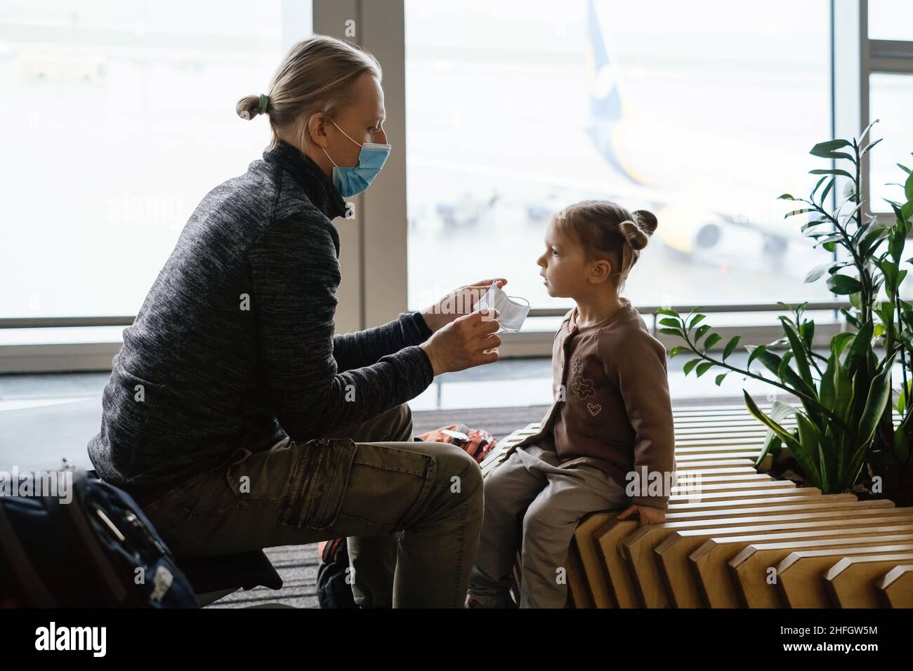 Padre che mette maschera sul viso dei bambini all'aeroporto. L'uomo protegge la ragazza dal coronavirus in luogo pubblico. Concetto di viaggio sicuro in famiglia. Foto Stock