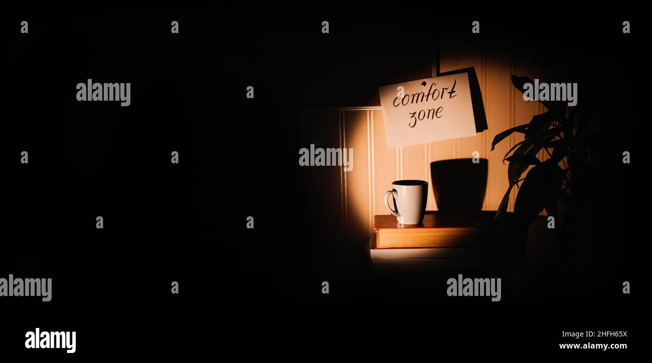 Concetto di zona comfort. Modi per uscire dalla zona comfort. Accogliente luogo a casa con tazza e libro in cerchio di luce in mezzo al buio Foto Stock