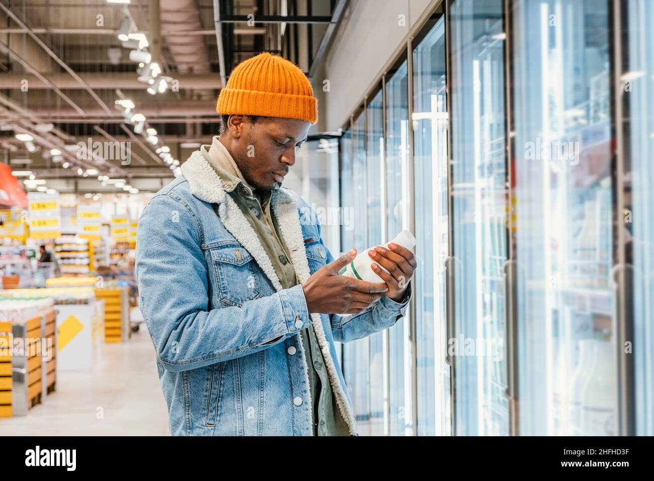 Il ragazzo afro-americano positivo in giacca di denim e cappello arancione prende la bottiglia di latte dalla scatola del frigorifero in vista laterale del moderno supermercato Foto Stock