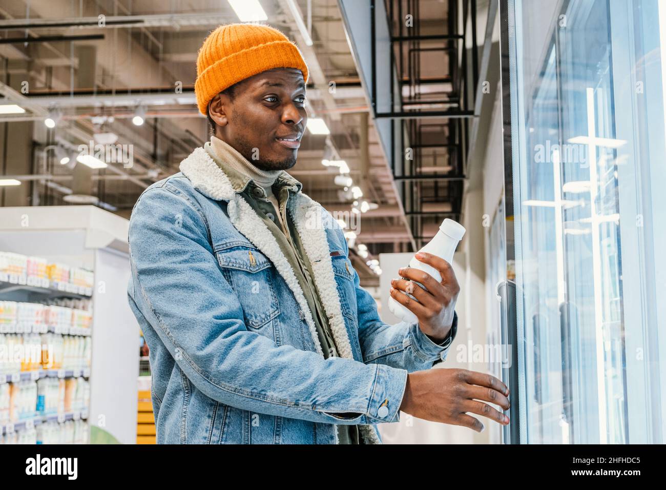 Il ragazzo afro-americano positivo in giacca di denim e cappello arancione prende la bottiglia di latte dalla scatola del frigorifero in vista laterale del moderno supermercato Foto Stock