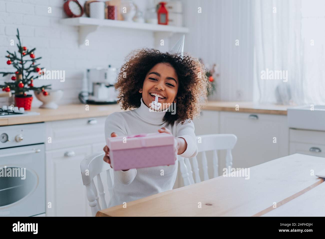 Sorridente signora africana con i capelli kinky tiene decorato presente scatola vicino al monitor del notebook seduta al primo piano della cucina moderna Foto Stock