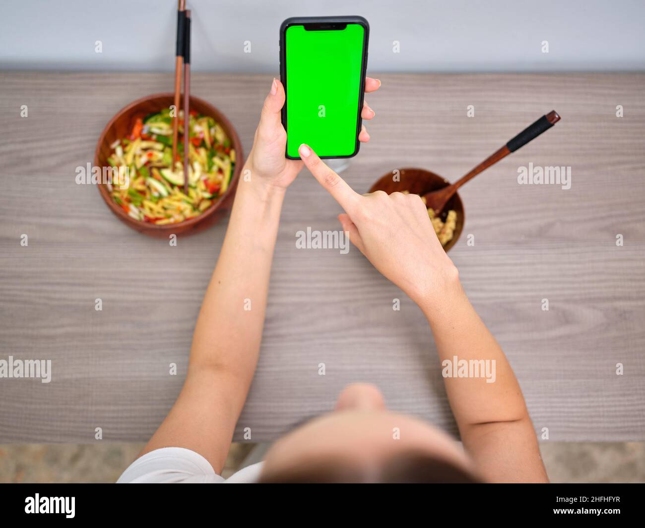 donna che mangia tabbouleh in una ciotola di legno con bacchette, guardando lo schermo verde del suo telefono cellulare Foto Stock