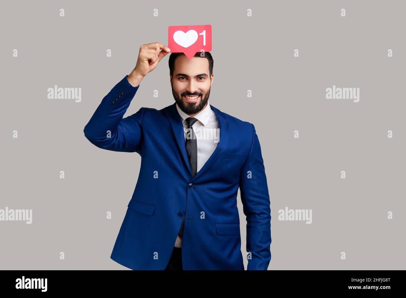 Uomo positivo che tiene il cuore come l'icona dei social media sopra la testa, pulsante di notifica per seguire, iscriversi blog, indossare abiti stile ufficiale. Studio interno girato isolato su sfondo grigio Foto Stock