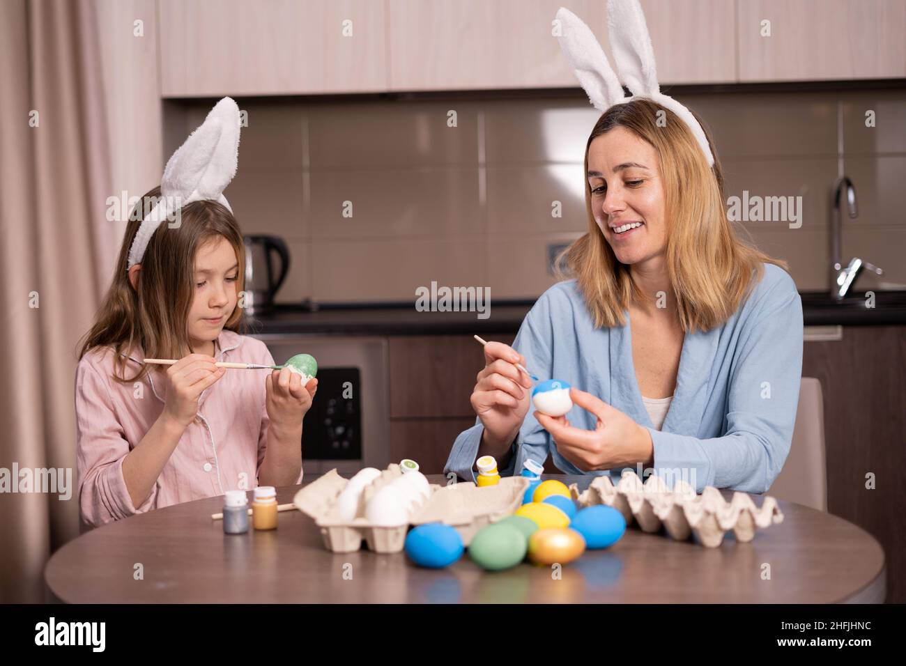 Madre e figlia dipingono le uova di Pasqua a casa in cucina. Buona famiglia si prepara per la Pasqua. Ragazza piccola carina con orecchie conigliate. Preparazione per Foto Stock