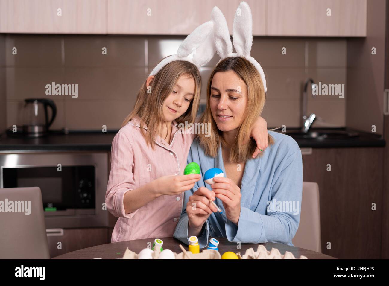 Madre e figlia dipingono le uova di Pasqua a casa in cucina. Buona famiglia si prepara per la Pasqua. Ragazza piccola carina con orecchie conigliate. Preparazione per Foto Stock