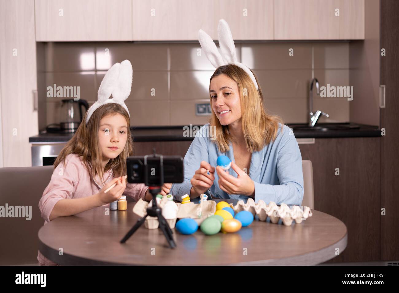 madre e figlia in un bendaggio sulle orecchie di un coniglio sono seduti in cucina a casa. Decorano le uova di Pasqua con le pitture e prendono selfie sopra Foto Stock