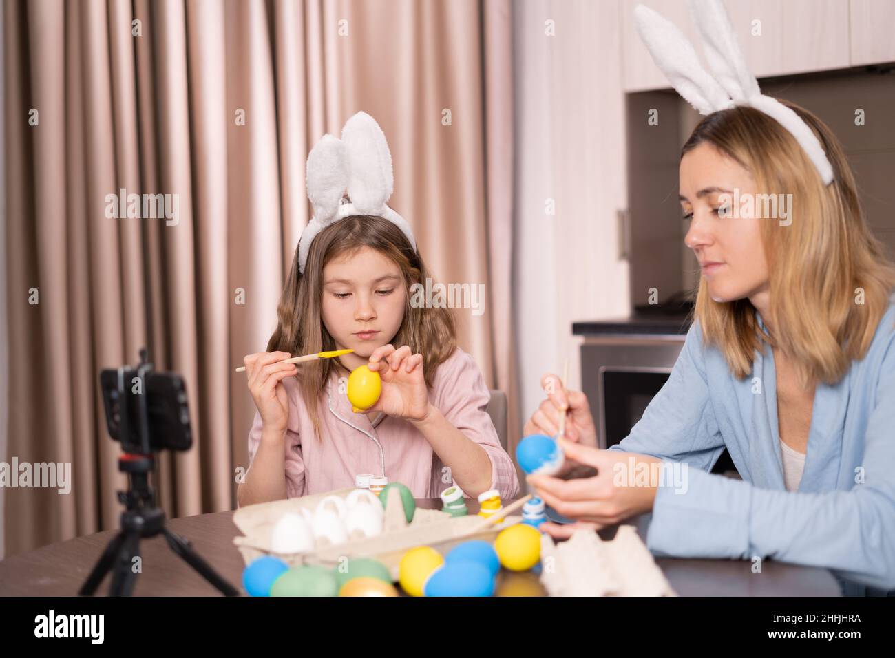 Madre caucasica e figlia in un bendaggio sulle orecchie di un coniglio sono seduti in cucina a casa. Decorare le uova di Pasqua con vernici e videochiamata Foto Stock