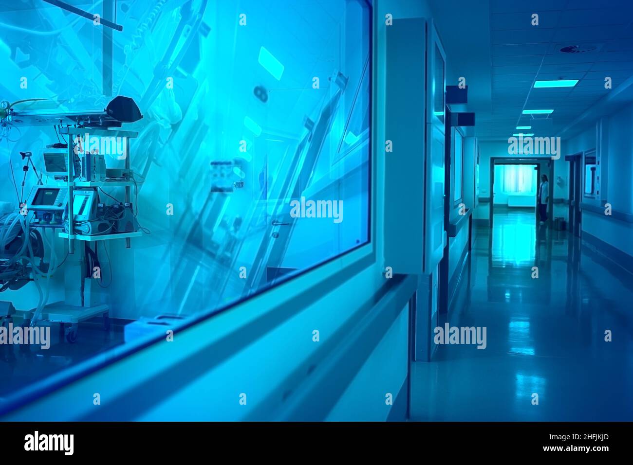 Corridoio dell'ospedale con finestra di osservazione al reparto con apparecchiatura riflettente e silhouette di medico femminile in fondo al corridoio. Foto Stock