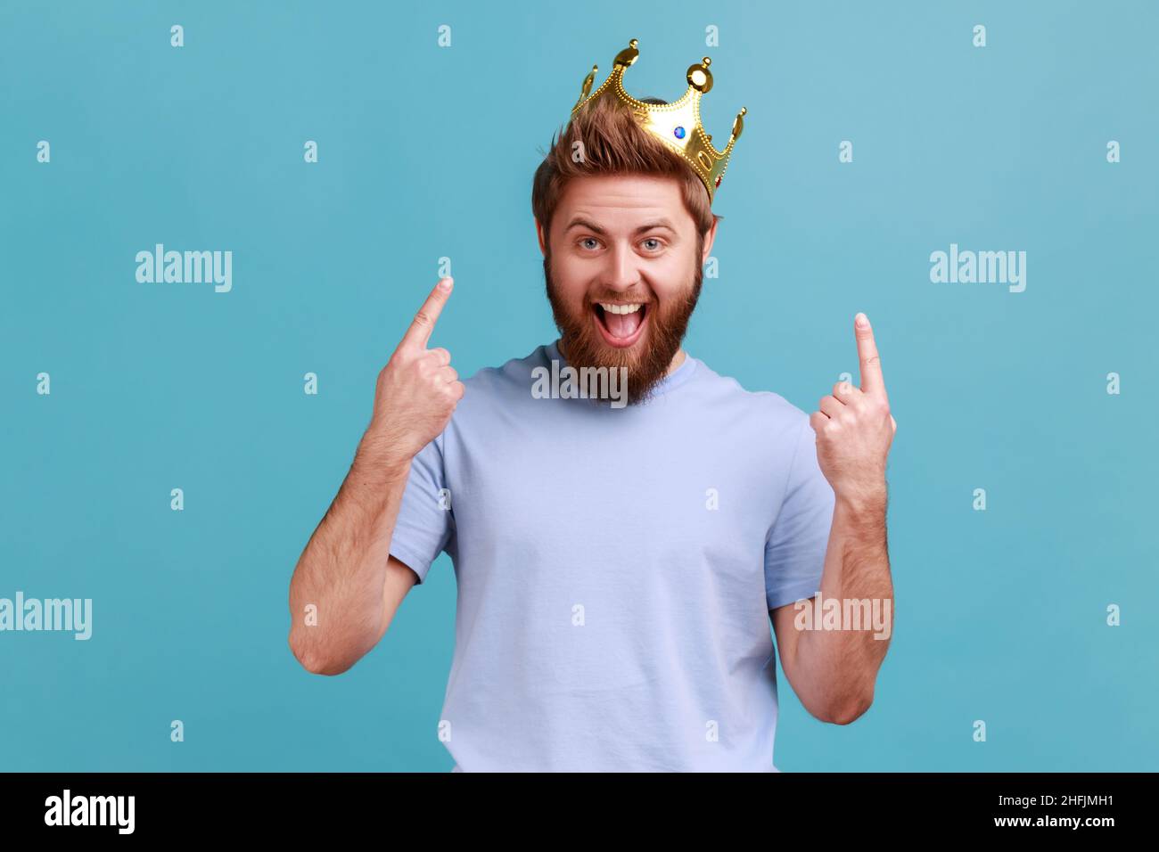 Ritratto di eccitato stupito uomo bearded puntando le dita sulla corona d'oro sulla sua testa, mostrando le sue qualità di leadership, concetto di autorità. Studio interno girato isolato su sfondo blu. Foto Stock