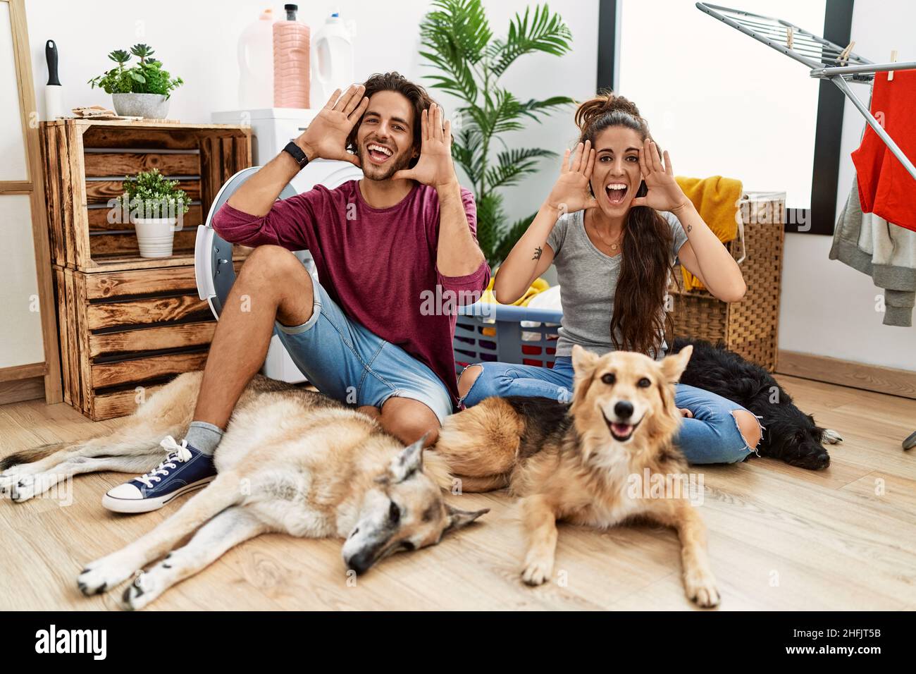 Giovane coppia ispanica che fa lavanderia con cani sorridenti allegri che giocano un boo con le mani che mostrano il volto. Sorpreso ed uscito Foto Stock