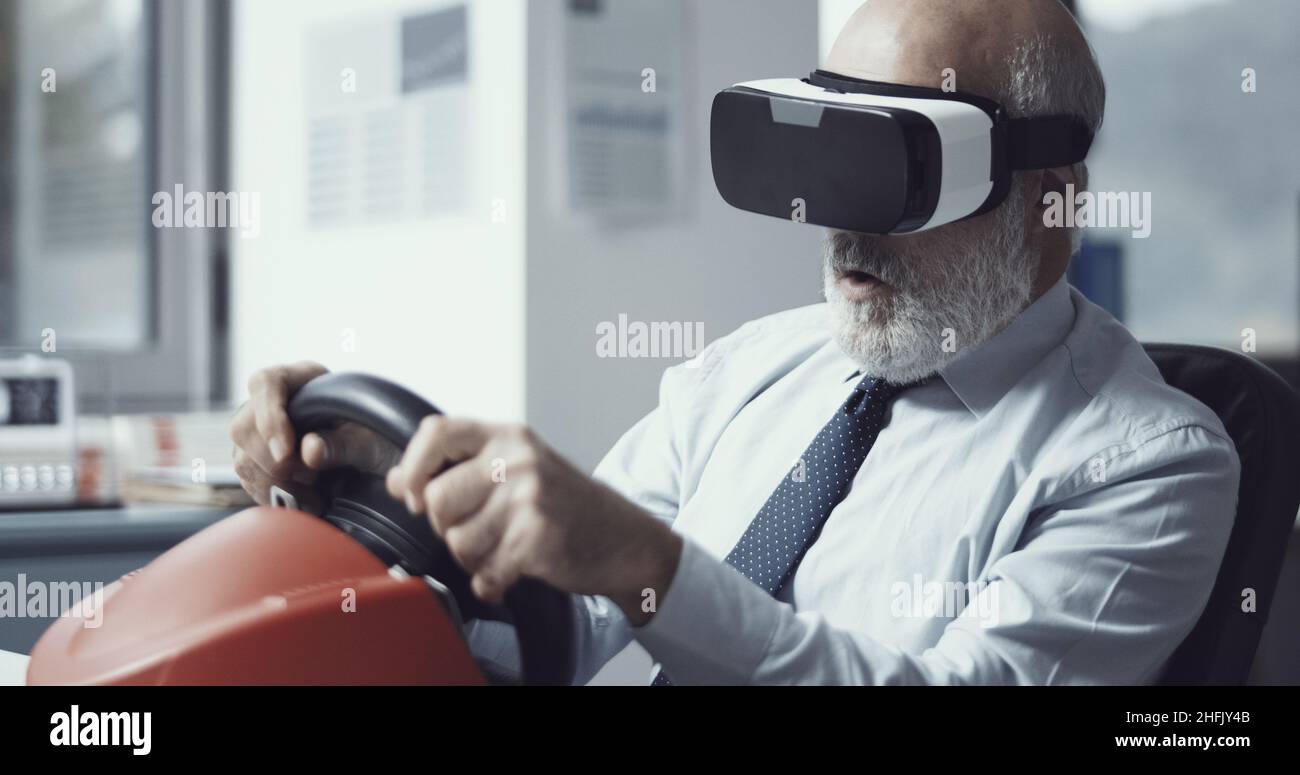 Un uomo d'affari felice che indossa un visore VR e gioca con una ruota da corsa in ufficio Foto Stock