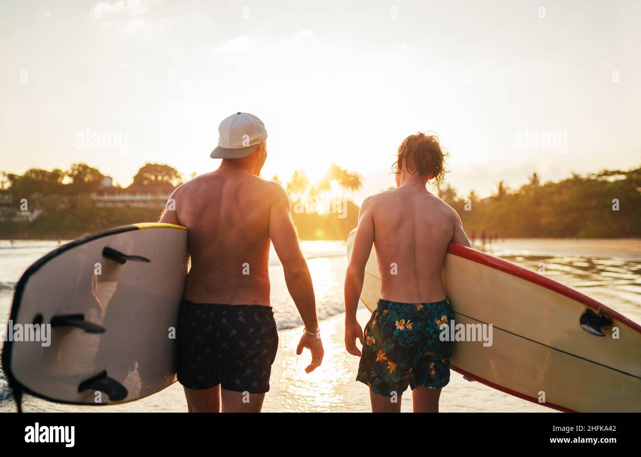 Padre con figlio adolescente che cammina con tavole da surf presso la spiaggia sabbiosa dell'oceano con palme sullo sfondo illuminate dal sole al tramonto. Sorridevano ed ha Foto Stock