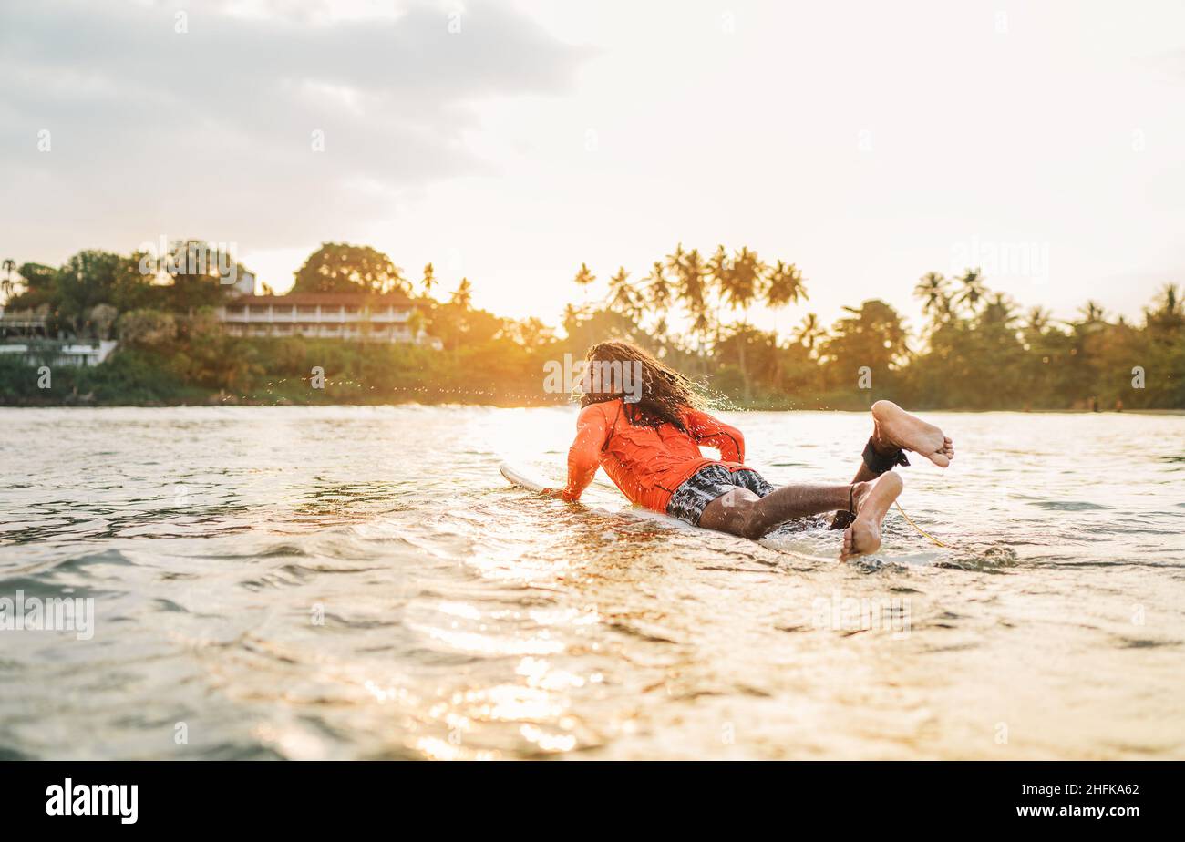 Uomo nero dai capelli lunghi che si paddling su una lunga tavola da surf al punto di surf nell'oceano Indiano. Palmeto ha littato raggi del tramonto sullo sfondo. Acqua estrema s Foto Stock