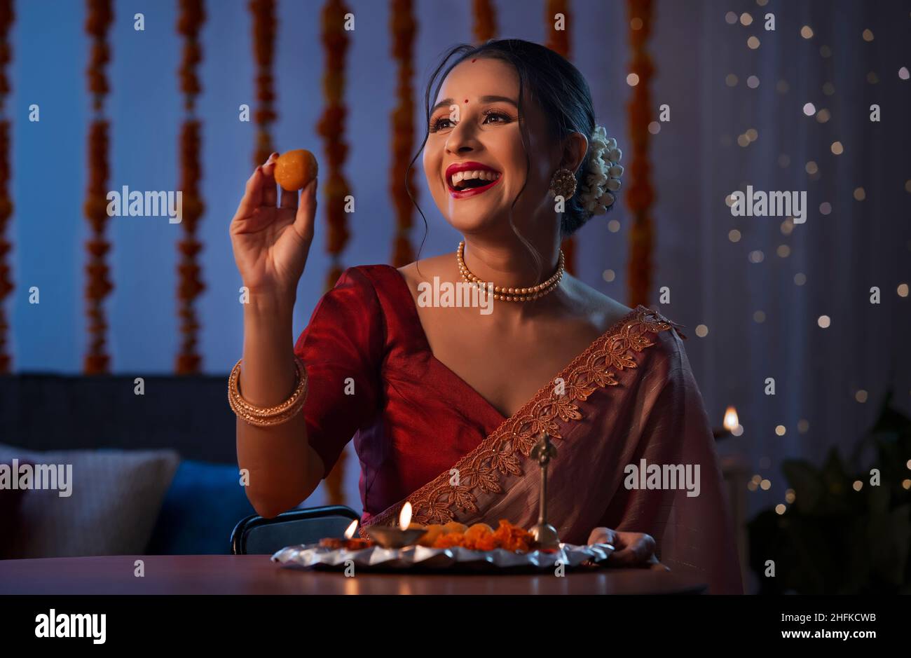 Una bella donna che tiene un laddoo e sorridente in mezzo dibali decorazione, luci e pooja thali. Foto Stock