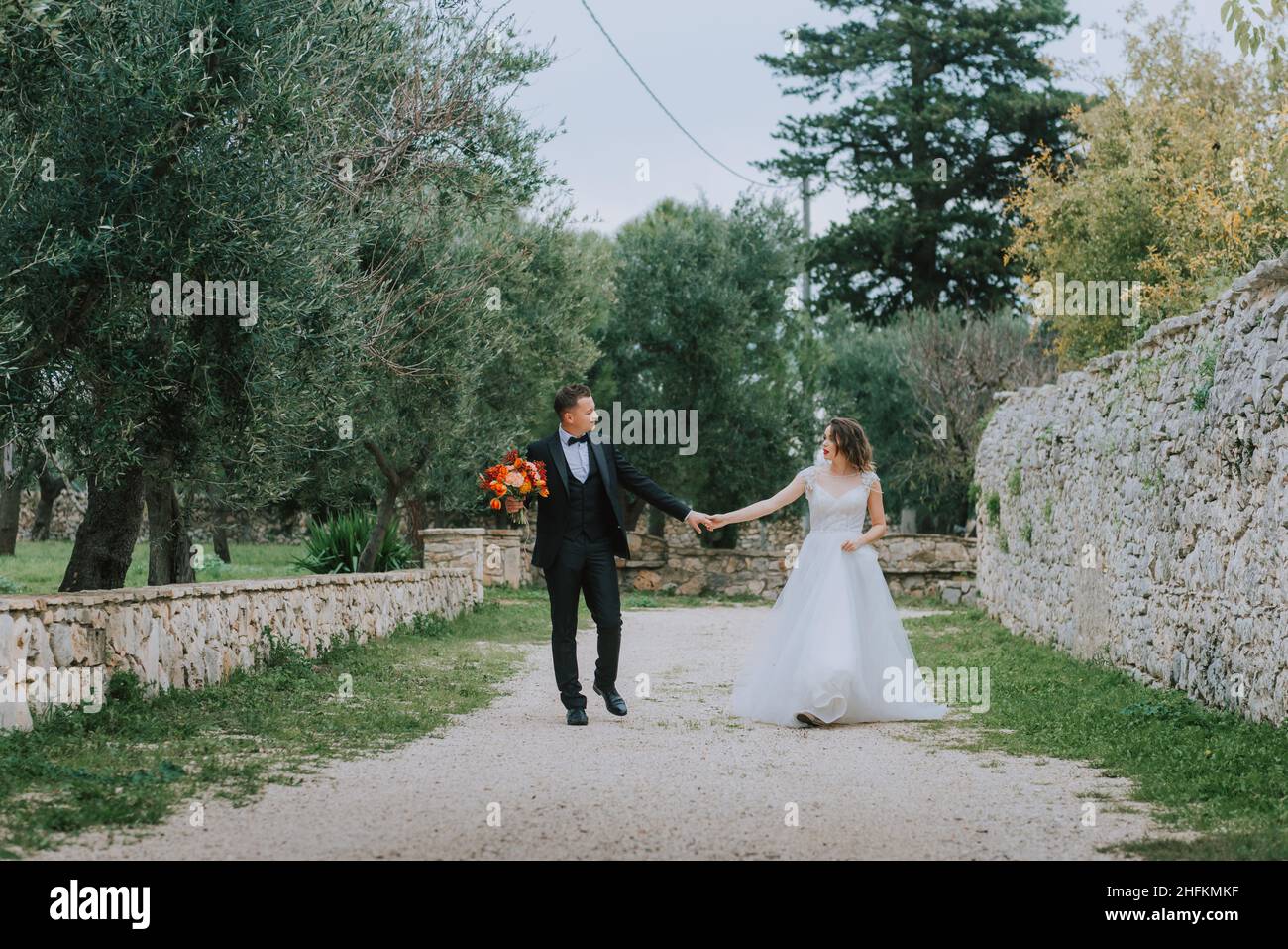 Felice coppia sorridente di stile che cammina in Toscana, Italia il giorno del matrimonio. La sposa e lo sposo camminano lungo la strada per le mani. Un giovane di stile Foto Stock