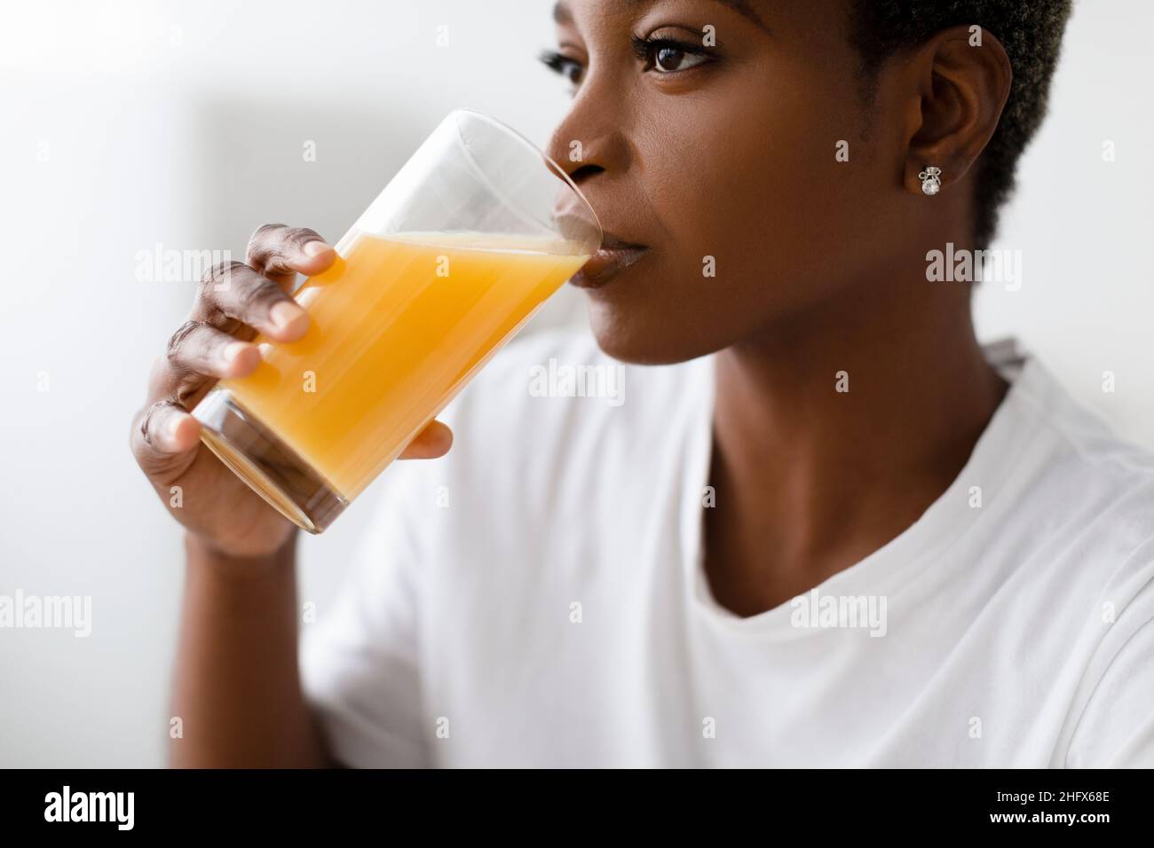 Cropped sete giovane graziosa signora nera in t-shirt bianco bevande succo d'arancia fresco da vetro interno, profilo Foto Stock