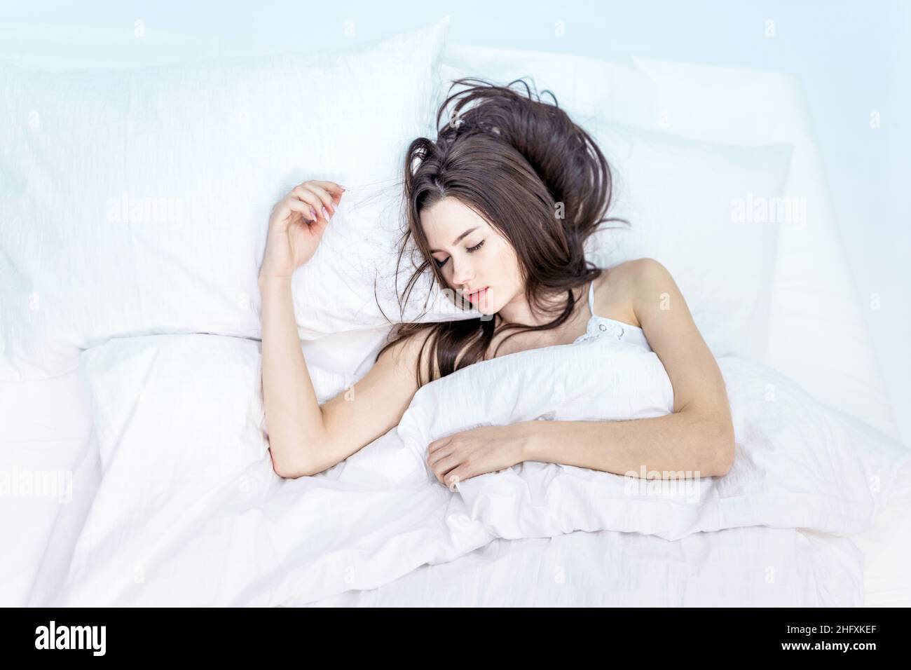 Buongiorno. Dormi a sufficienza. Camera da letto. La brunetta dorme su un letto bianco. Vedere sogni piacevoli. Una ragazza carina in pigiama si trova sul letto con la sua e. Foto Stock