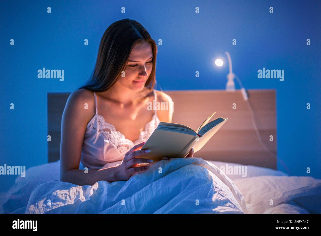 Una bella donna legge un libro con luci soffuse nella sua camera da letto prima di andare a letto. Relax. Letteratura. Autosviluppo Foto Stock