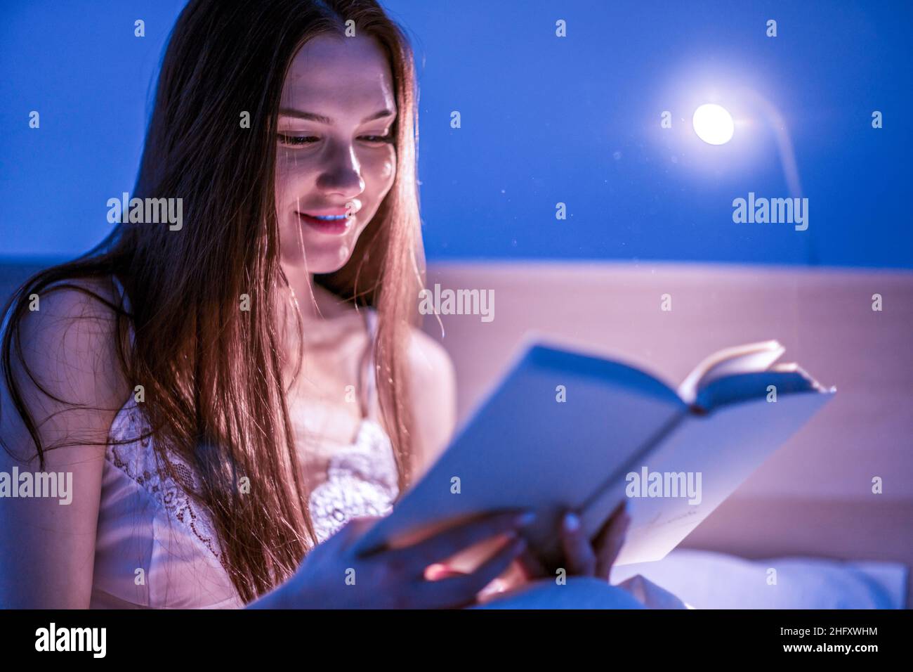 Una bella donna legge un libro con luci soffuse nella sua camera da letto prima di andare a letto. Autosviluppo. Letteratura. Relax. Foto Stock