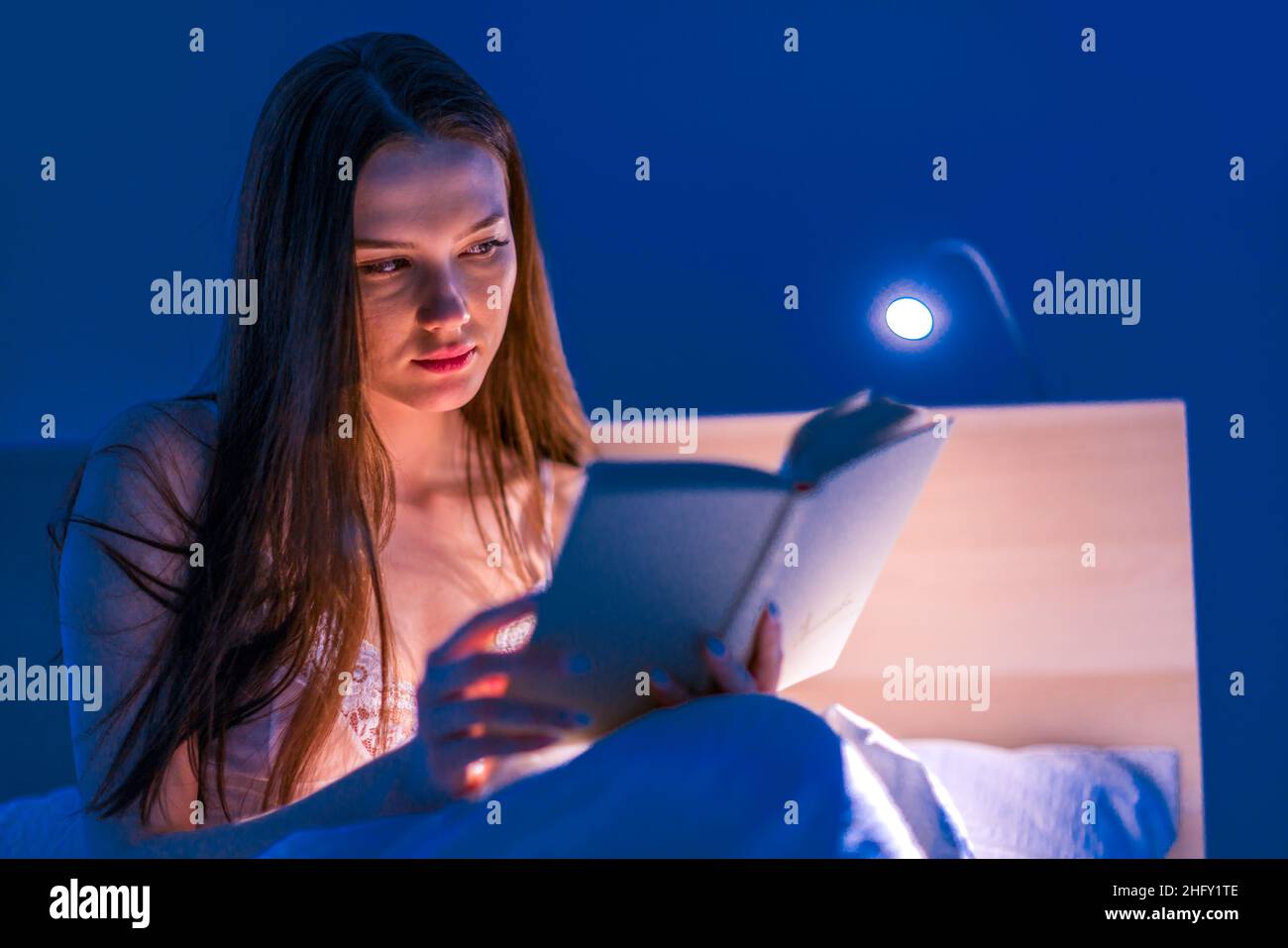 Una bella donna legge un libro con luci soffuse nella sua camera da letto prima di andare a letto. Letteratura. Autosviluppo. Relax. Foto Stock