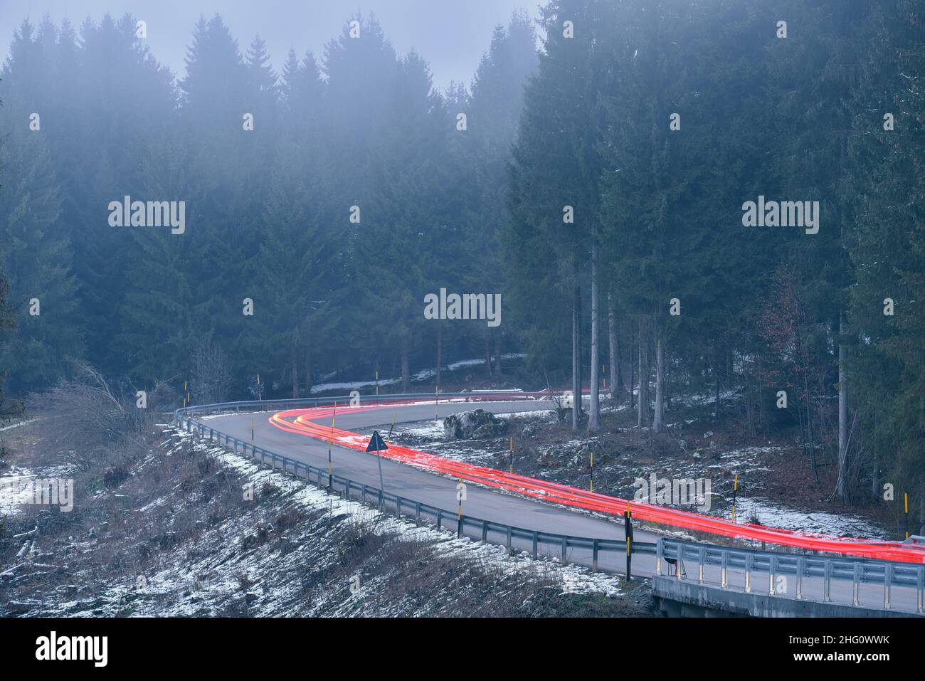 Percorsi leggeri dalle luci di coda su una tortuosa strada alpina attraverso una pineta in una notte d'inverno nebbia Foto Stock