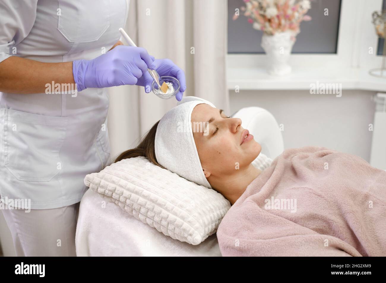 Il cosmetologo applica la maschera della pelle con pennello, cura e procedura di trattamento in clinica estetica moderna. Donna tratta la pelle in salone di bellezza, anti-età non Foto Stock