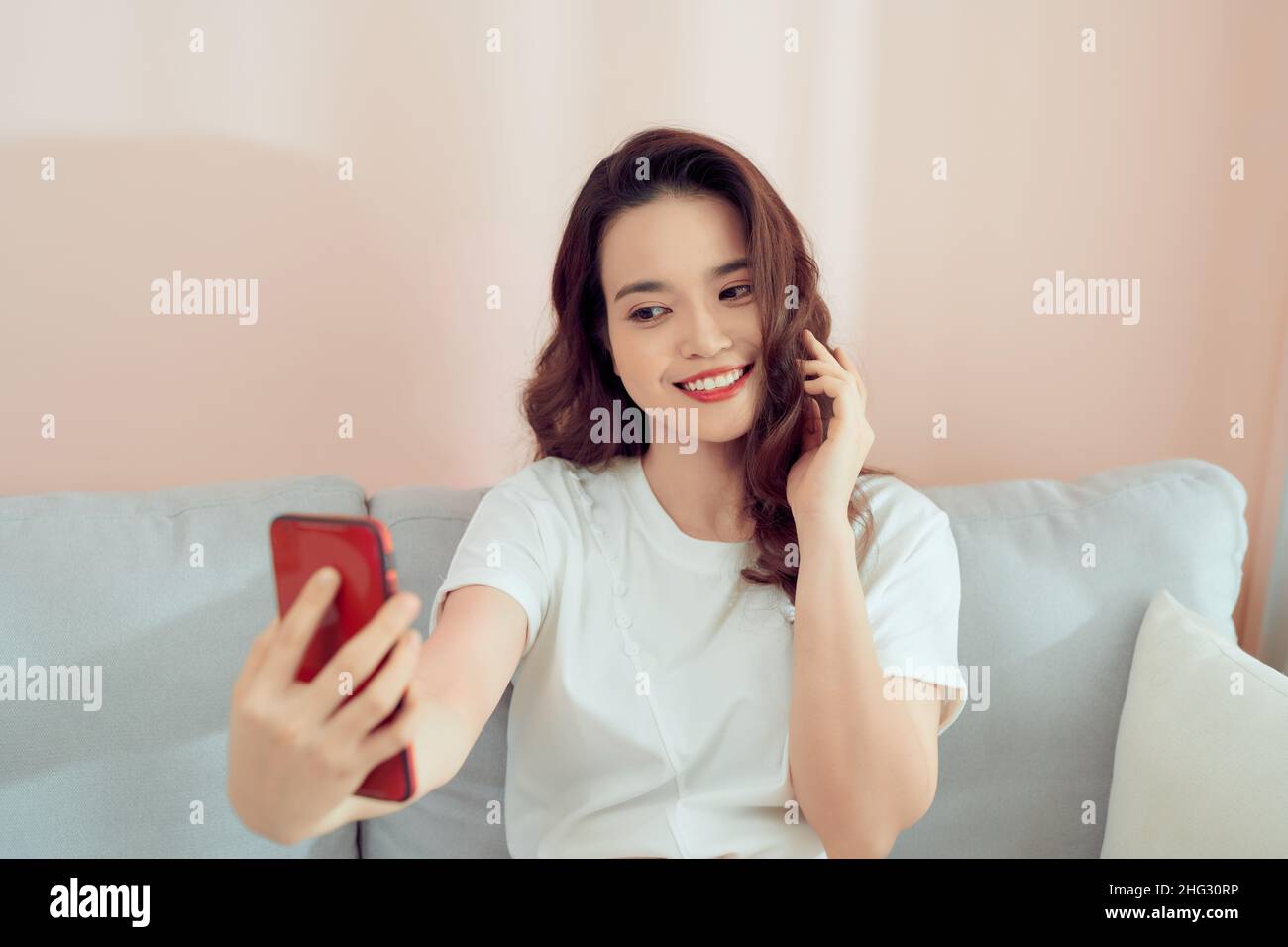 Scatto di una giovane donna asiatica felice che prende selfie con il suo cellulare mentre si siede in soggiorno. Foto Stock