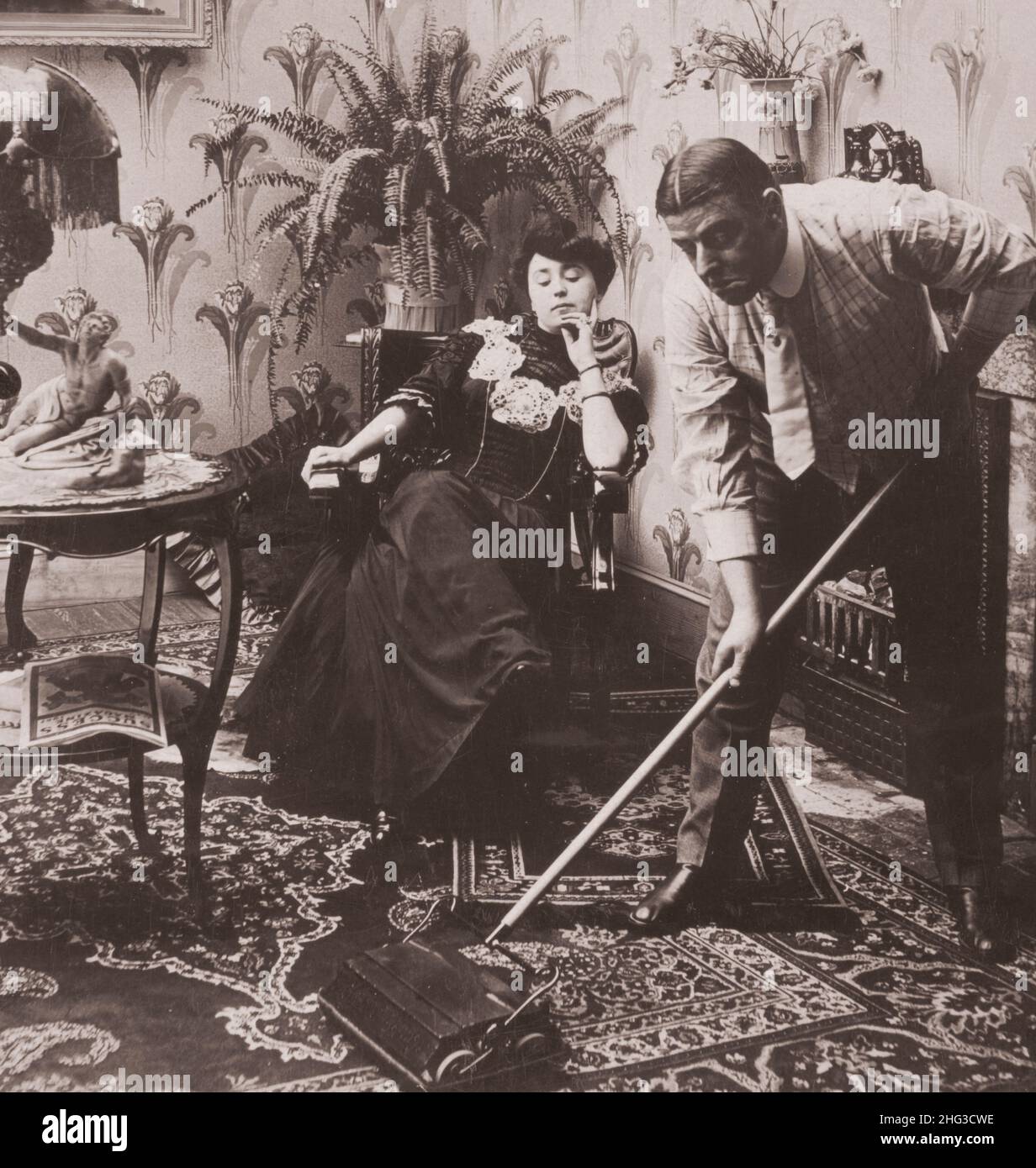 Vintage foto d'amore. 'Prenda il vostro tempo il sig. Peck- la fretta fa spreco '. USA. 1907 uomo che usa la spazzatrice da tappeto come donna si siede e guarda. Foto Stock