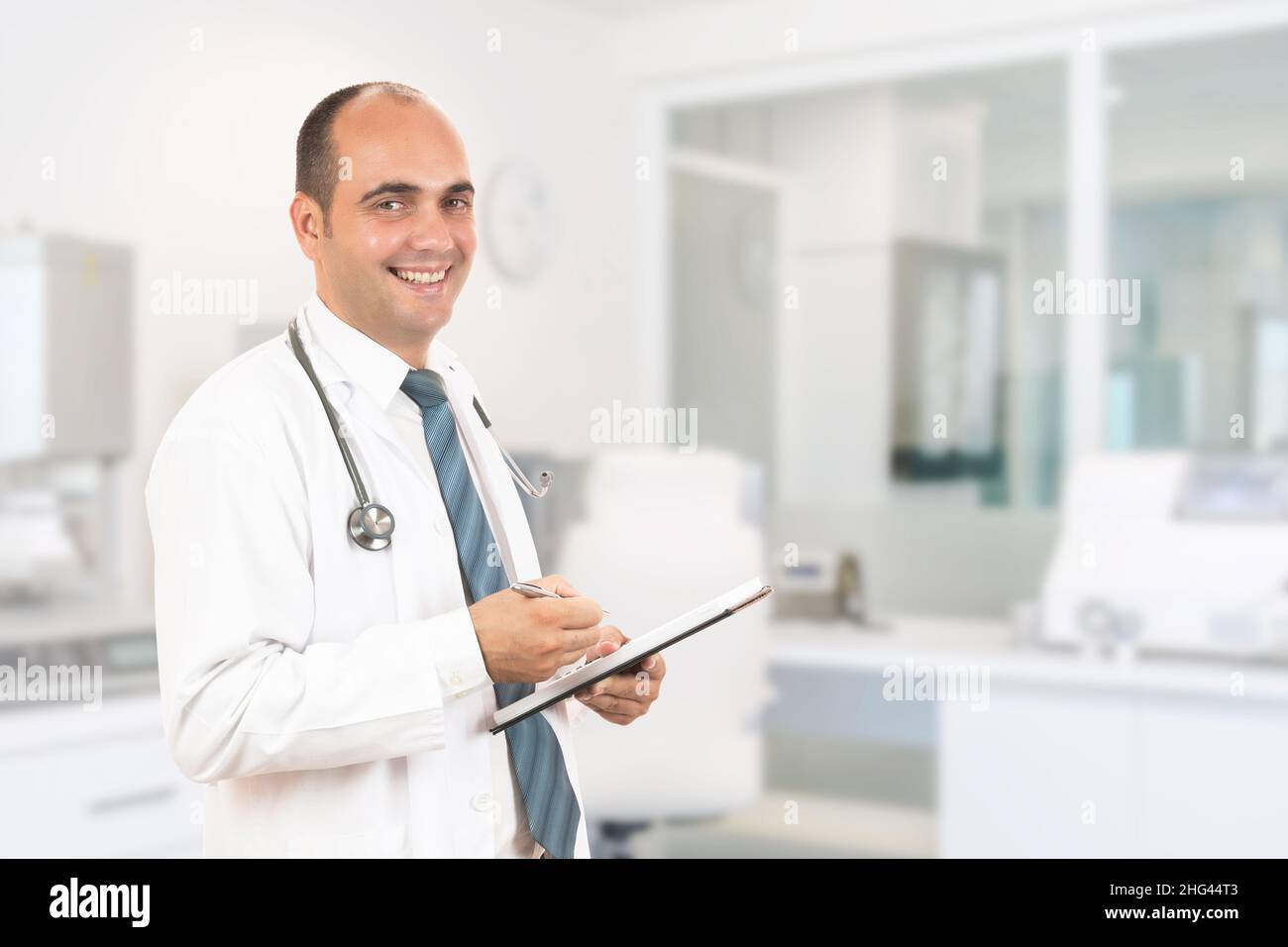 Medico maschile con stetoscopio che tiene un taccuino. Sorridente medico maschile che compila il modulo medico sulla clipboard mentre si alza dritto in ospedale Foto Stock