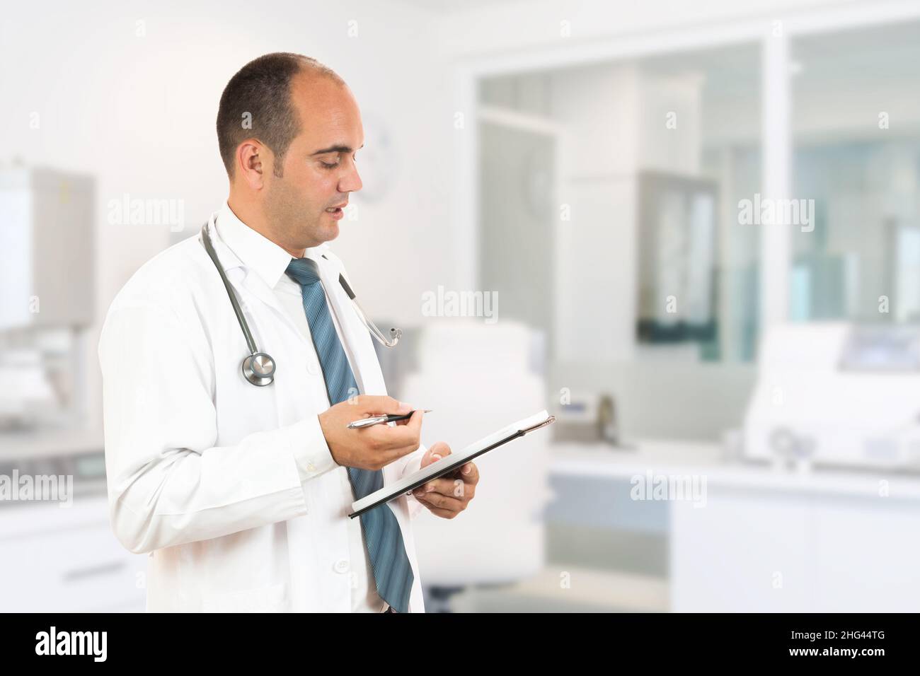 Medico maschile con stetoscopio che tiene un taccuino. Sorridente medico maschile che compila il modulo medico sulla clipboard mentre si alza dritto in ospedale Foto Stock
