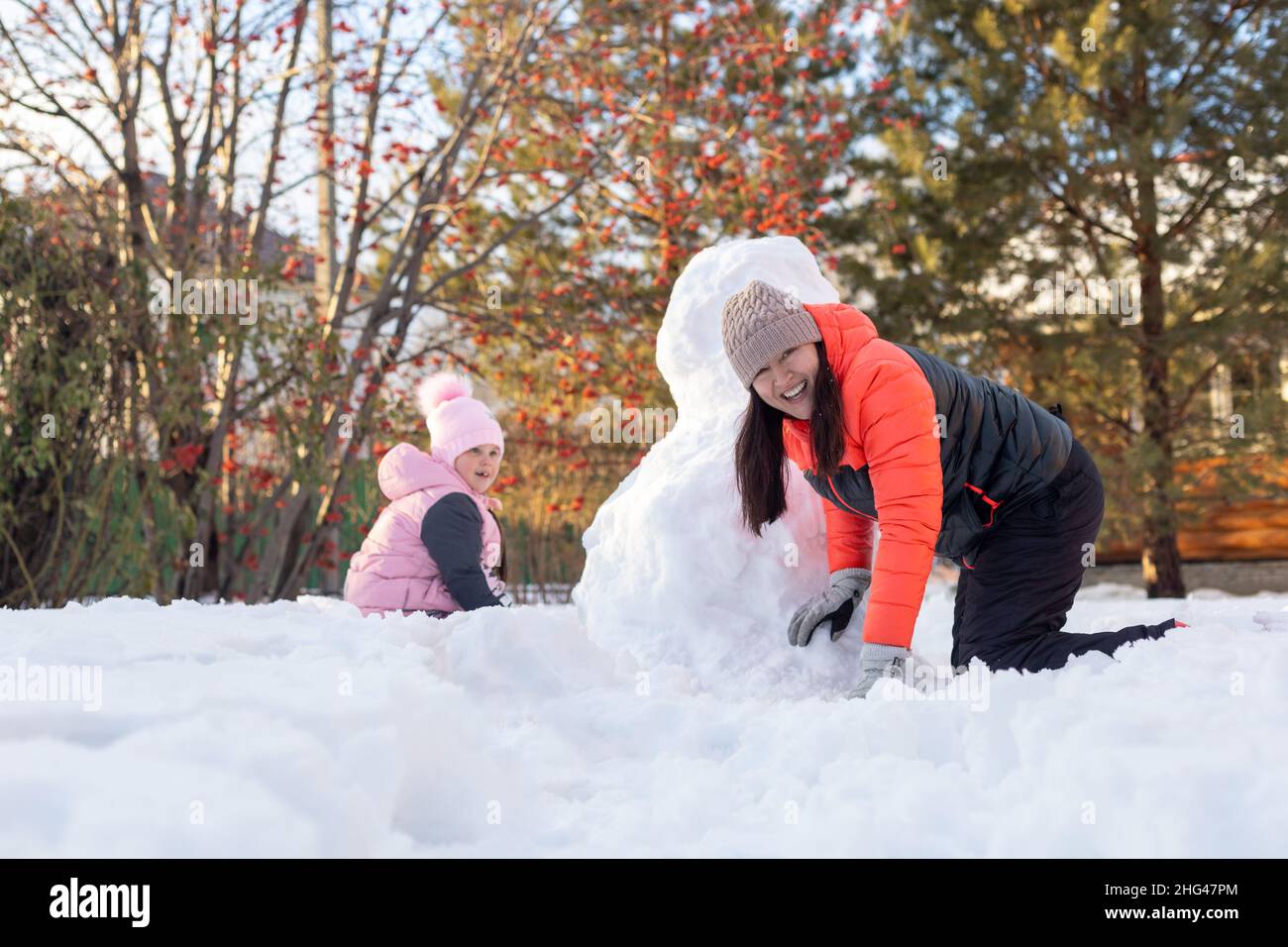 Ritratto di bambina e madre strisciando sulla neve vicino nevaio sul cortile in serata con rowan e abeti sullo sfondo. I genitori trascorrono del tempo Foto Stock