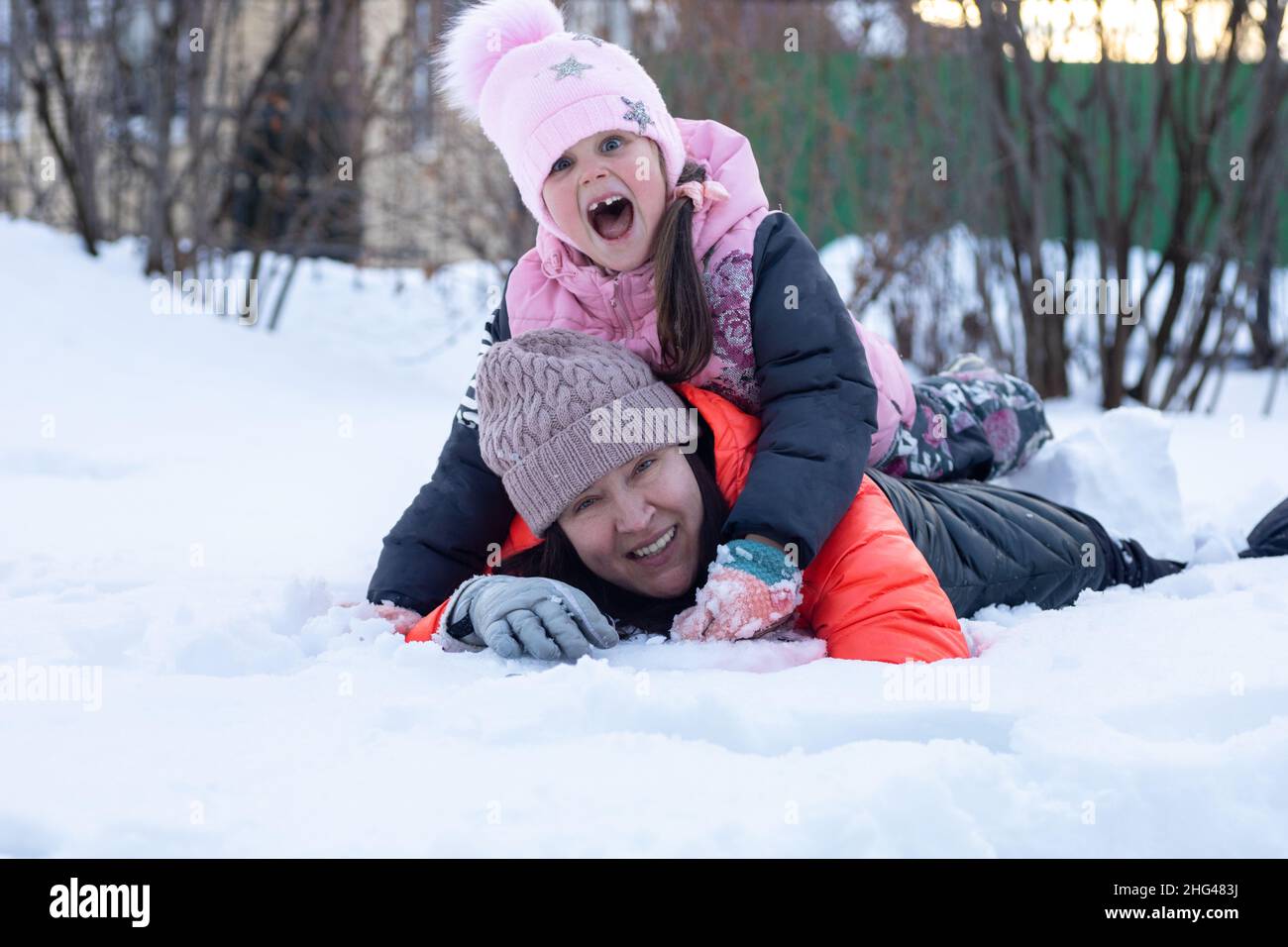Madre di mezza età con figlia sdraiata sulla neve insieme sorridendo con i denti nel parco di sera con gli alberi sullo sfondo. I genitori trascorrono del tempo Foto Stock