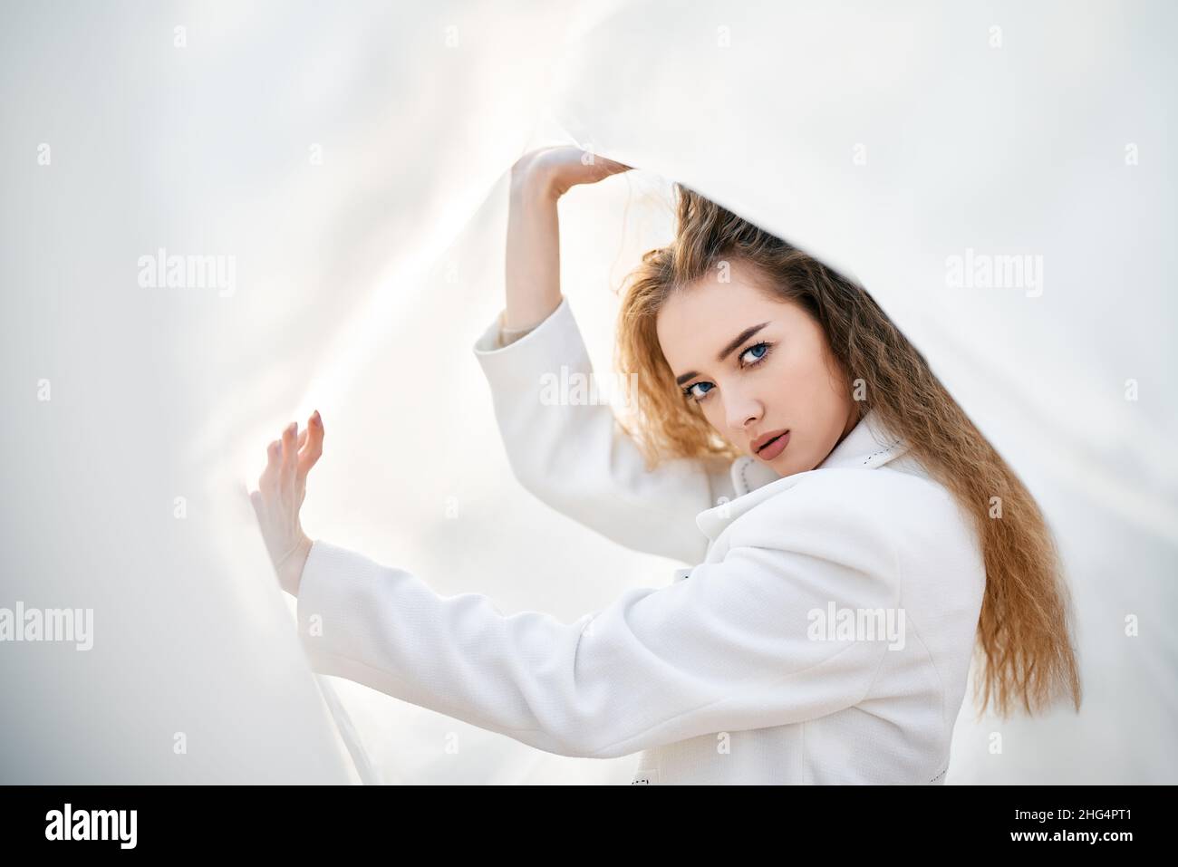 Giovane donna attraente in abiti bianchi in posa su sfondo astratto pallido guardando la macchina fotografica sicuro. Colori tenui e soleggiati. Concetto di bellezza Foto Stock
