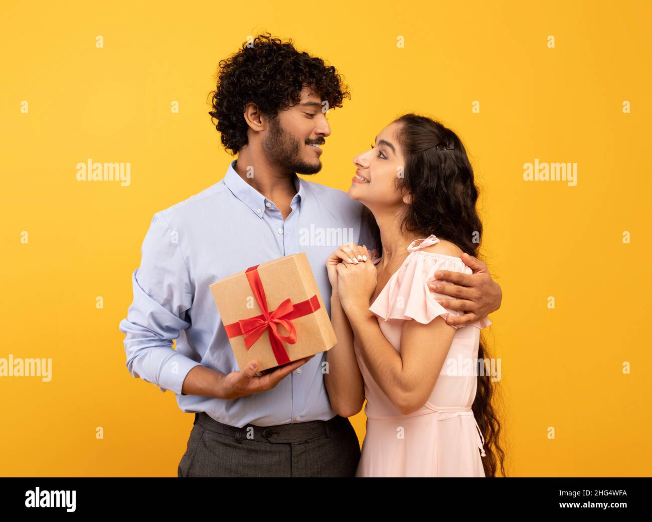 Concetto di celebrazione. Amorevole coppia indiana abbracciando e l'uomo che tiene avvolto scatola regalo, sfondo giallo studio Foto Stock