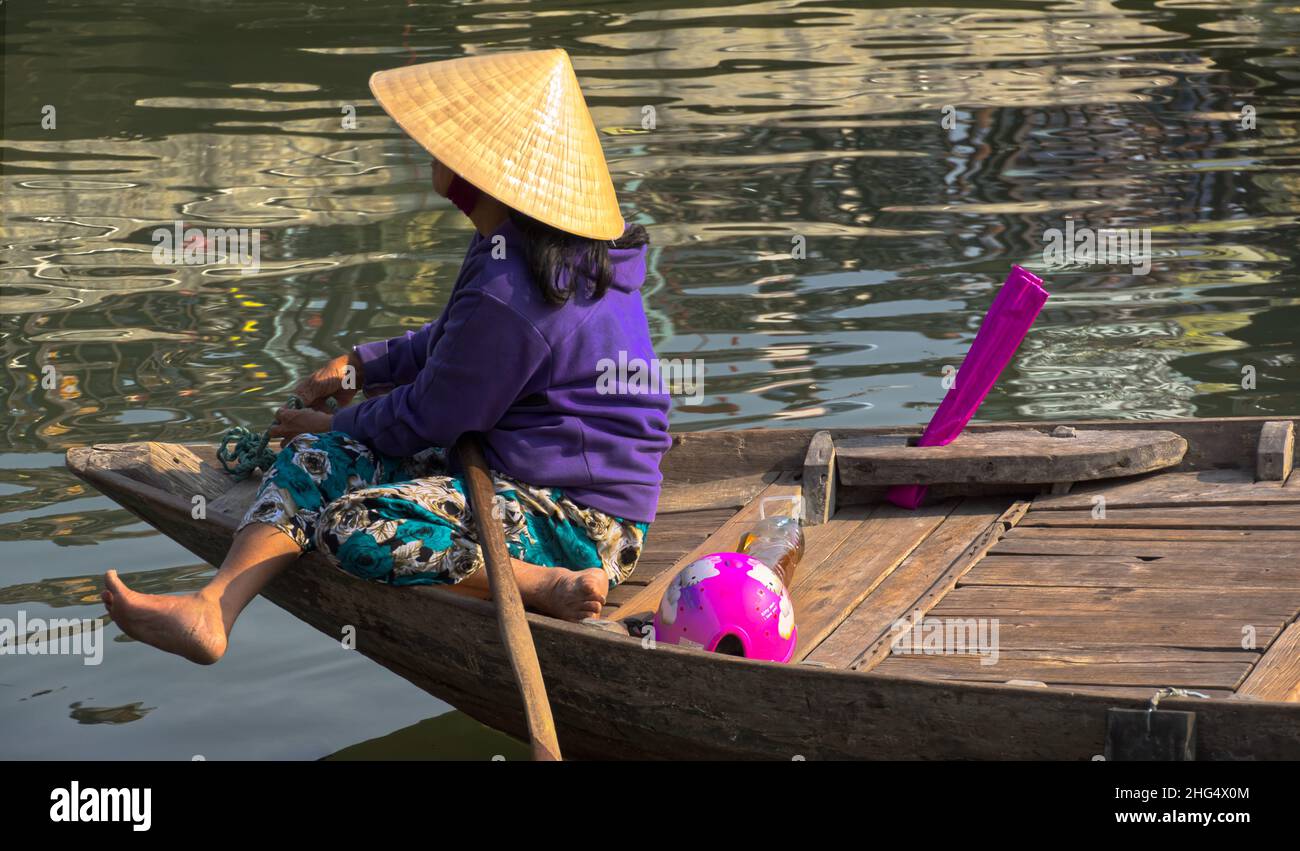 Eine junge Vietnamesin mit Reis- bzw. Kegelhut sitzt vorne auf ihrem kleinen Holzboot und blickt auf den Song-Thu-Bon Fluss in Hoi An Foto Stock