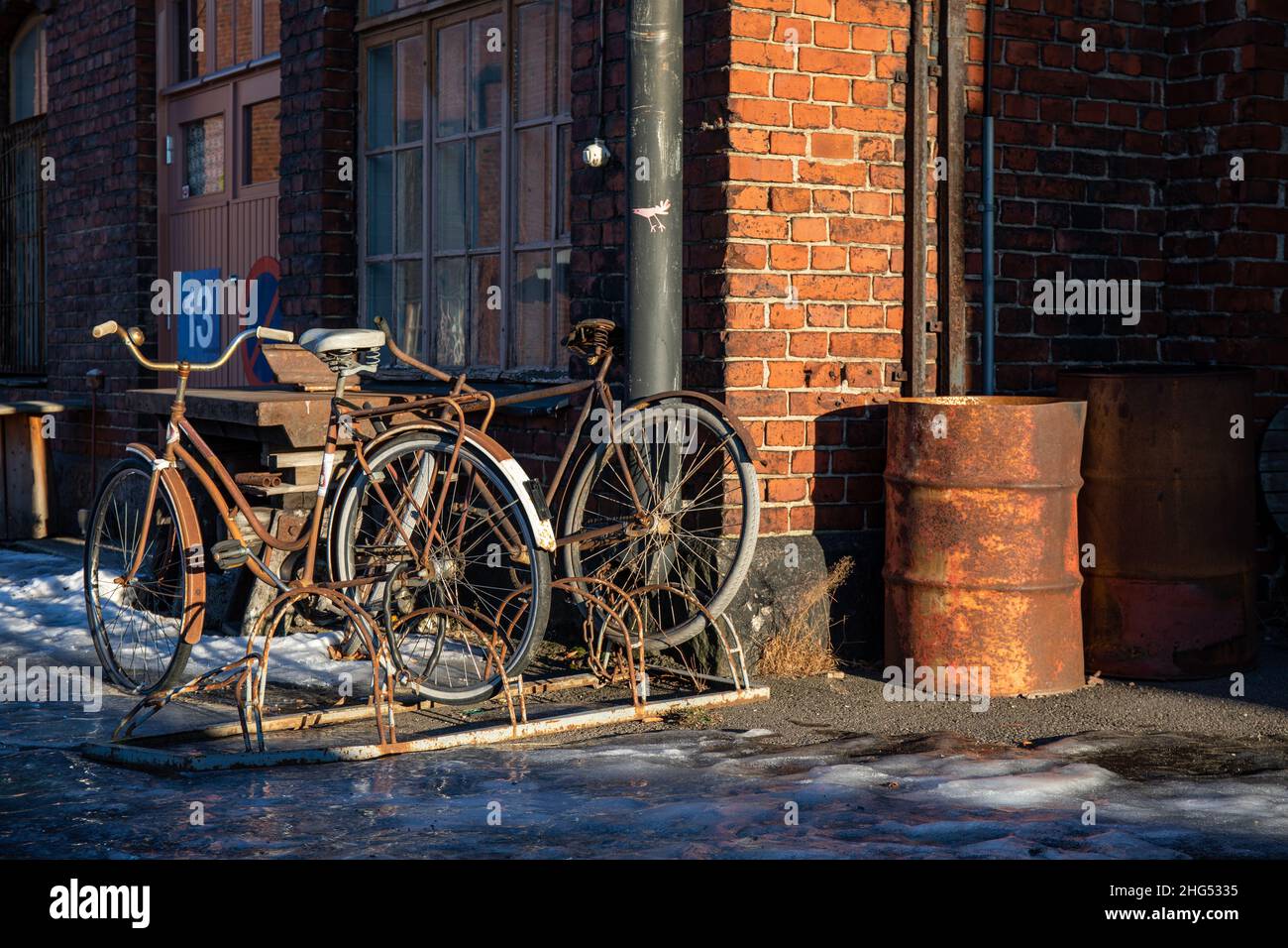 Biciclette arrugginite e tamburo di petrolio contro vecchio edificio di mattoni rossi nel sole del pomeriggio sul vecchio cantiere ferroviario di Pasila, Helsinki, Finlandia Foto Stock
