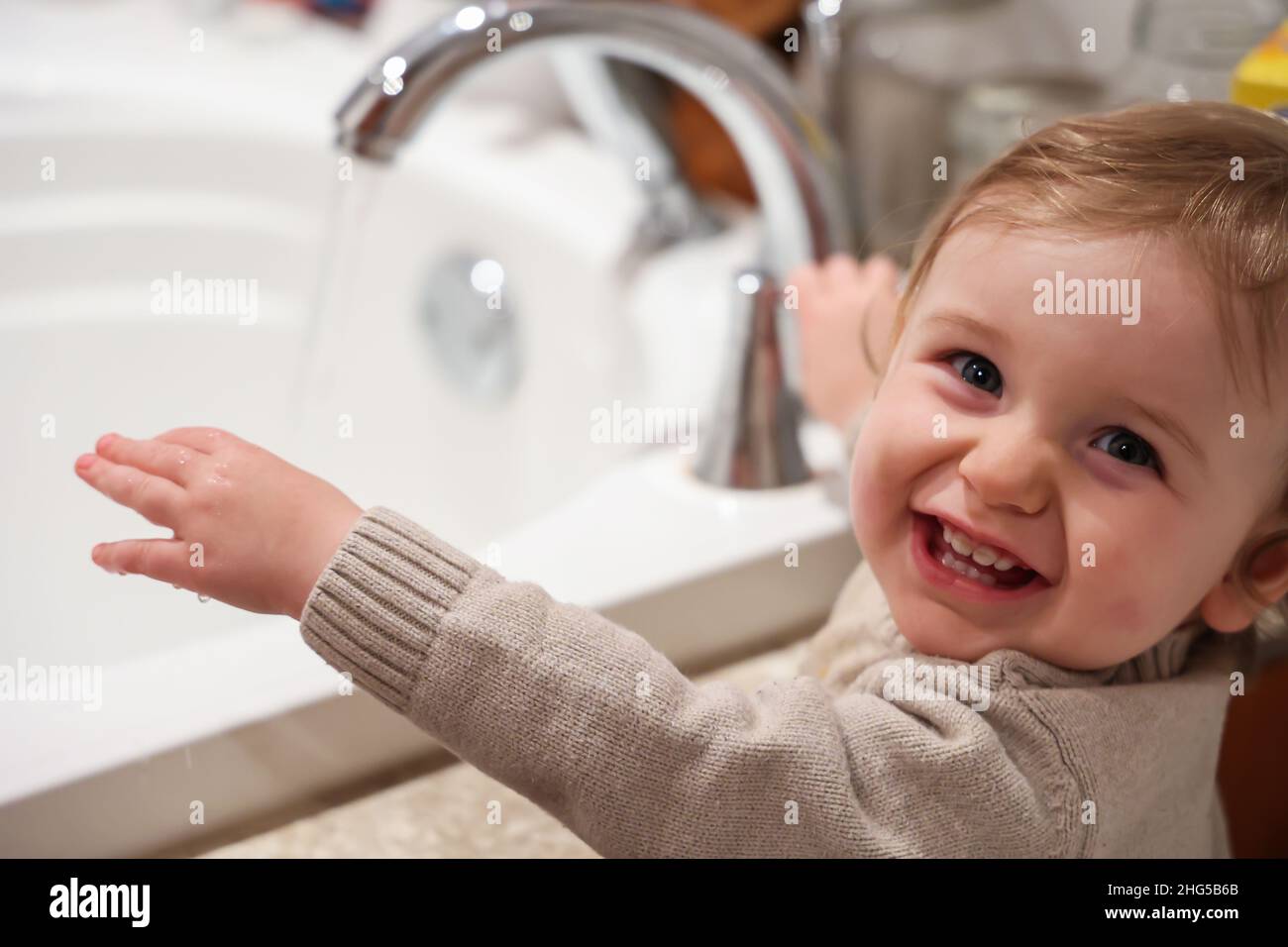 Un ragazzo gioioso di tre anni è visto da vicino in bagno in fuoco selettivo. Lavando le mani con il bacino sfocato e l'acqua corrente corrente sullo sfondo. Foto Stock