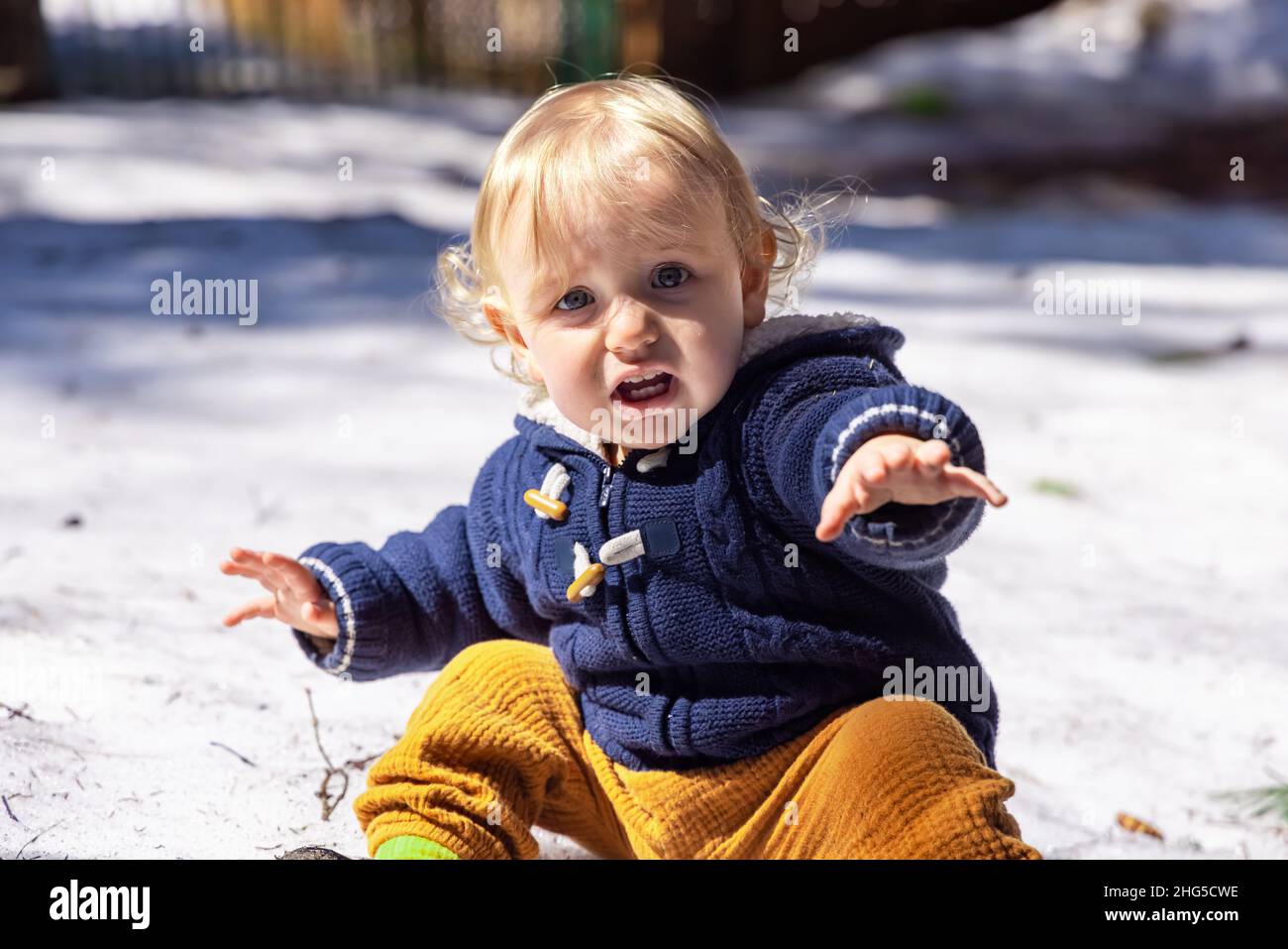 Vista frontale di un ragazzo di 3 anni con espressione preoccupata, occhi spalancati e bocca aperta, raggiungendo la mano a macchina fotografica seduta sulla neve con sfondo sfocato. Foto Stock