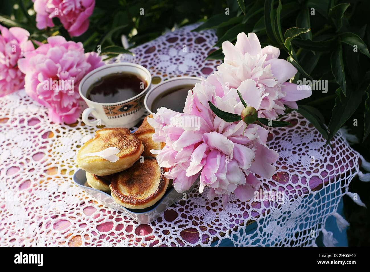 Tè in stile country nel giardino estivo nel villaggio. Due tazze di tè nero e pancake fatti a mano su tovaglie vintage crocheted e pe fiorente Foto Stock