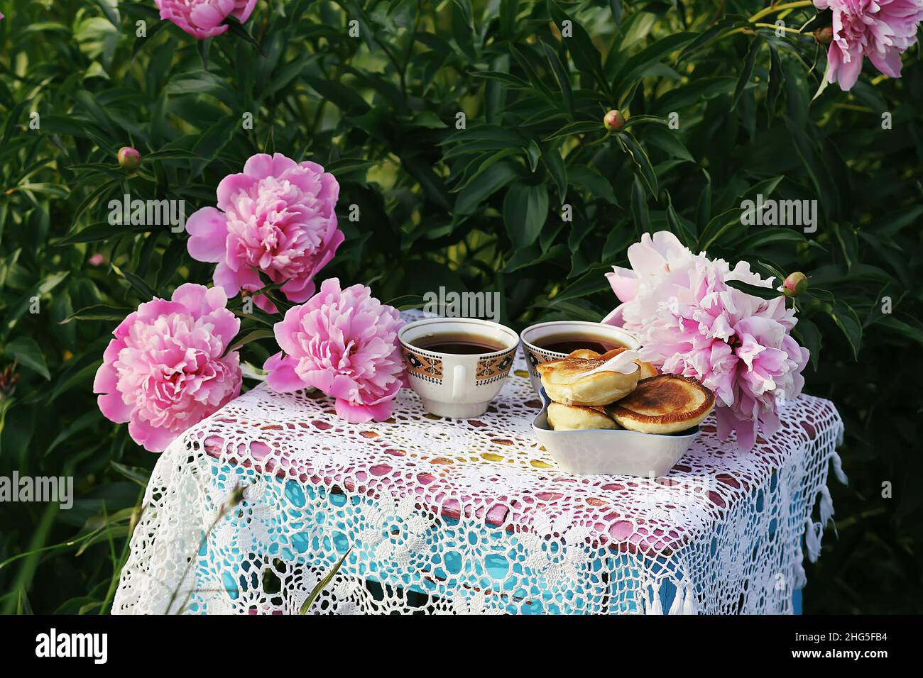 Tè in stile country nel giardino estivo nel villaggio. Due tazze di tè nero e pancake fatti a mano su tovaglie vintage crocheted e pe fiorente Foto Stock