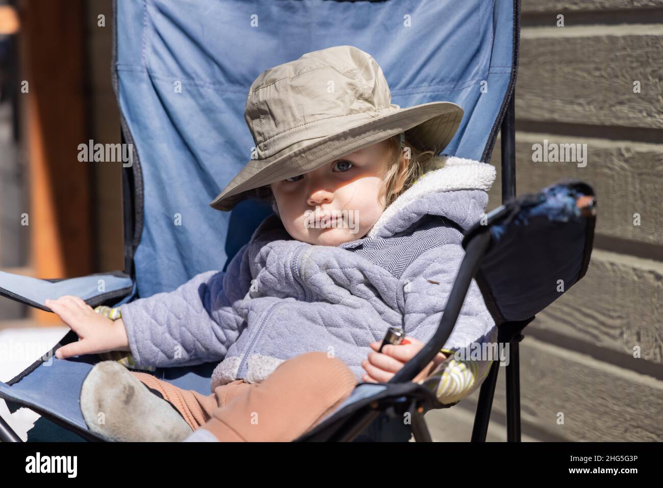 Ritratto a fuoco selettivo del toddler di 2 anni, seduto su una sedia da campeggio all'aperto che indossa un cappello a secchio brimed per mantenere il sole luminoso fuori dagli occhi gentili. Foto Stock