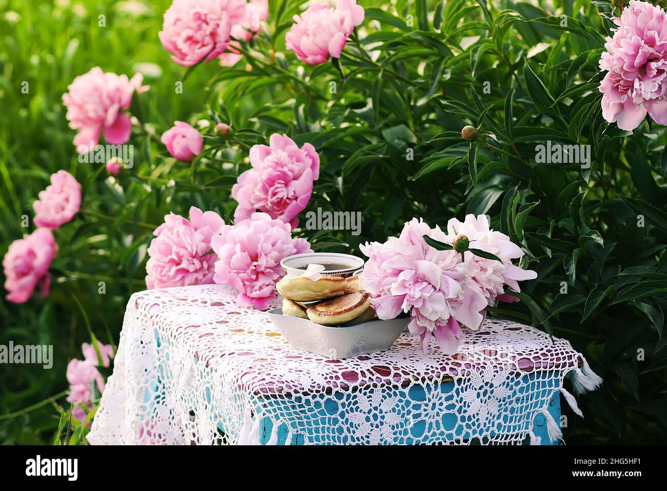 Tè in stile country nel giardino estivo nel villaggio. Due tazze di tè nero e pancake fatti a mano su tovaglie vintage crocheted e pe fiorente Foto Stock