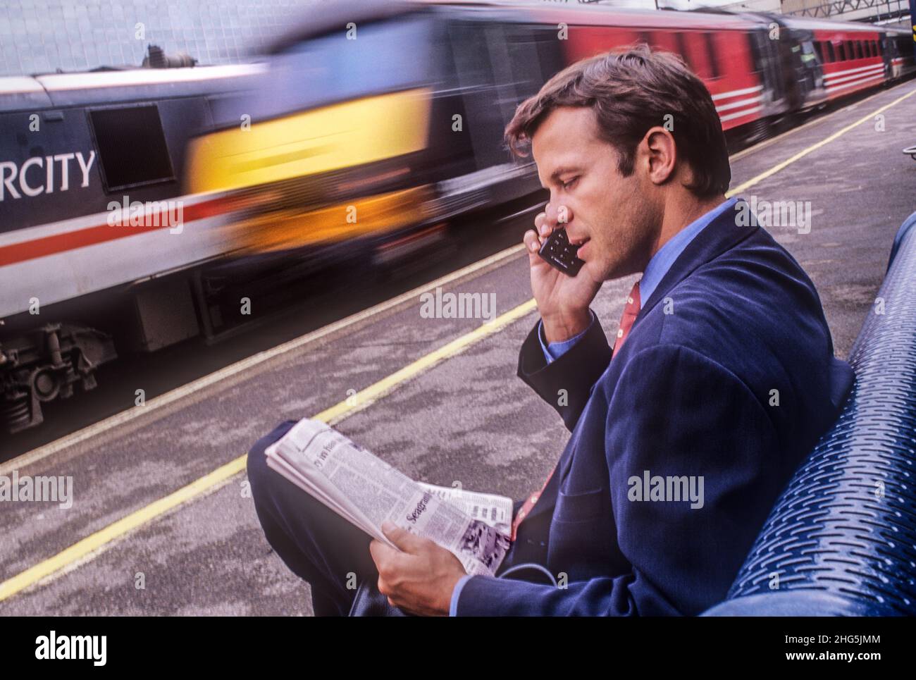 1990s retro Travel businessman London train blur, parlando su telefono cellulare seduto sul banco della stazione ferroviaria britannica. Lettura di un giornale e parlando sulla tecnologia più recente 90s piccolo telefono cellulare Ericsson. Intercity Cross Country Virgin treno di passaggio con sfocatura velocità. Moda stile tecnologia di viaggio e trasporto negli anni '1990s Foto Stock