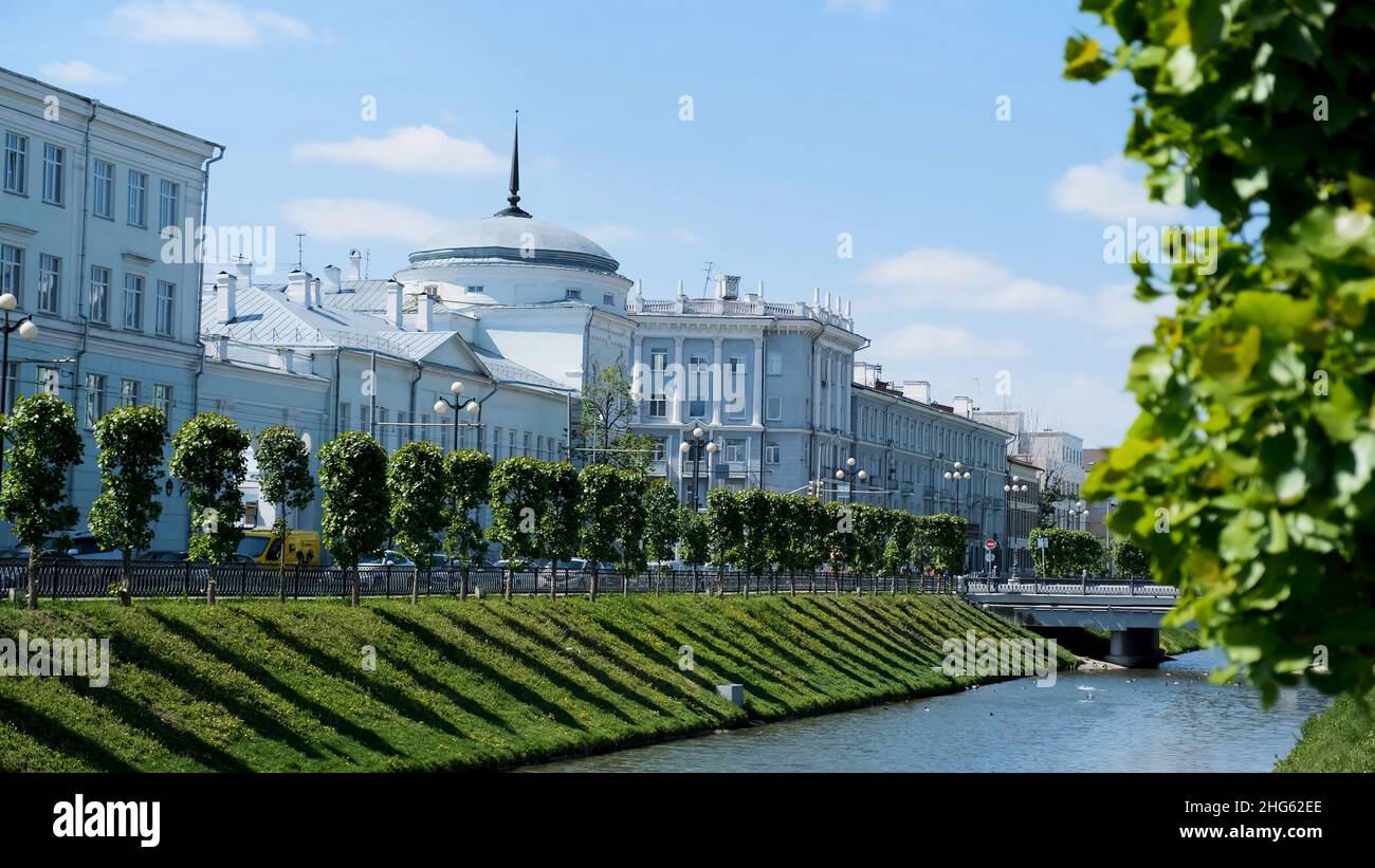 Bellissimo paesaggio urbano con un canale stretto e due pendii coperti da erba verde su sfondo blu cielo nuvoloso. Summer City Street con case bianche, e. Foto Stock